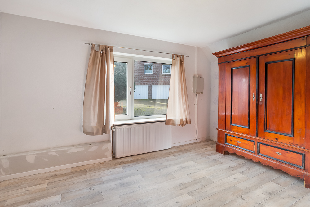 Zimmer mit Fenster, beigen Vorhängen und großem Holzschrank auf hellem Holzboden.