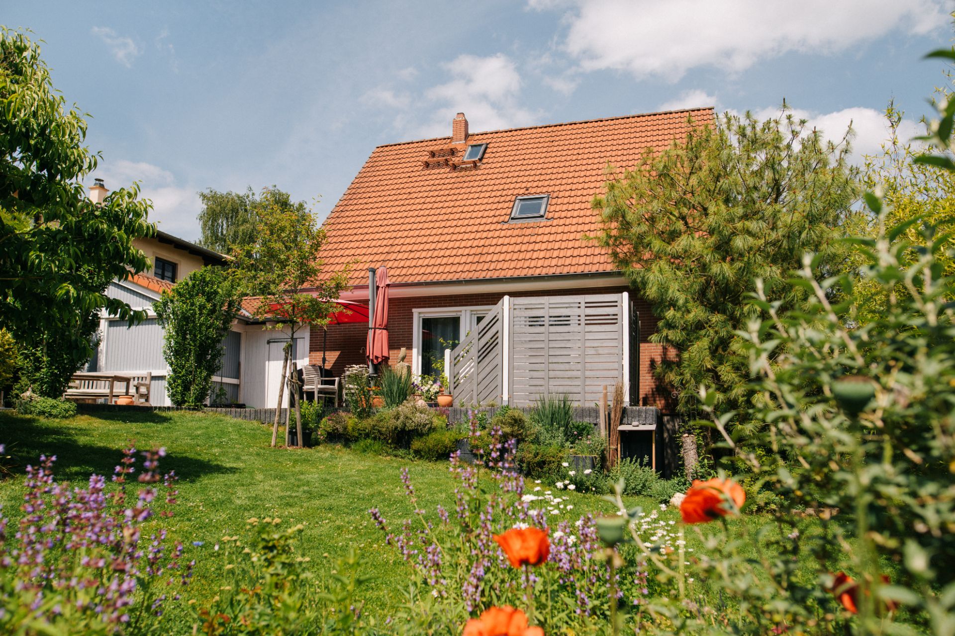 Haus mit rotem Dach und Garten, Blumen und Terrasse mit Sonnenschirm im Vordergrund.