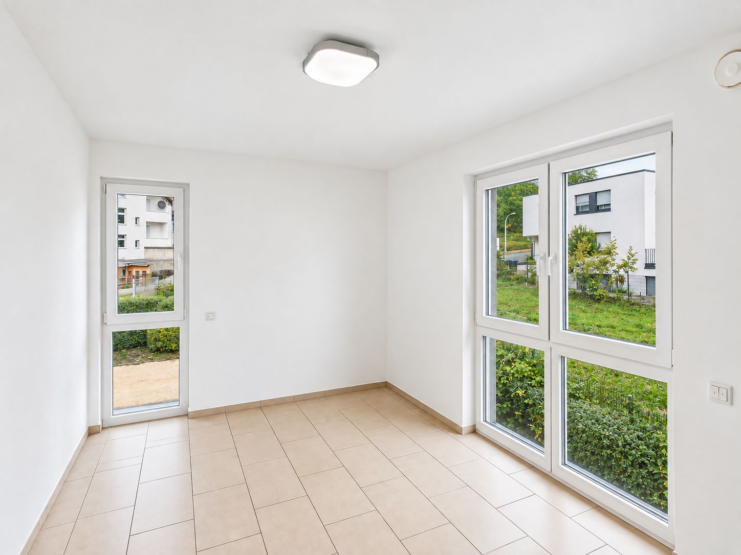 Heller Raum mit weißen Wänden, Fliesenboden und großen Fenstern mit Blick ins Grüne.