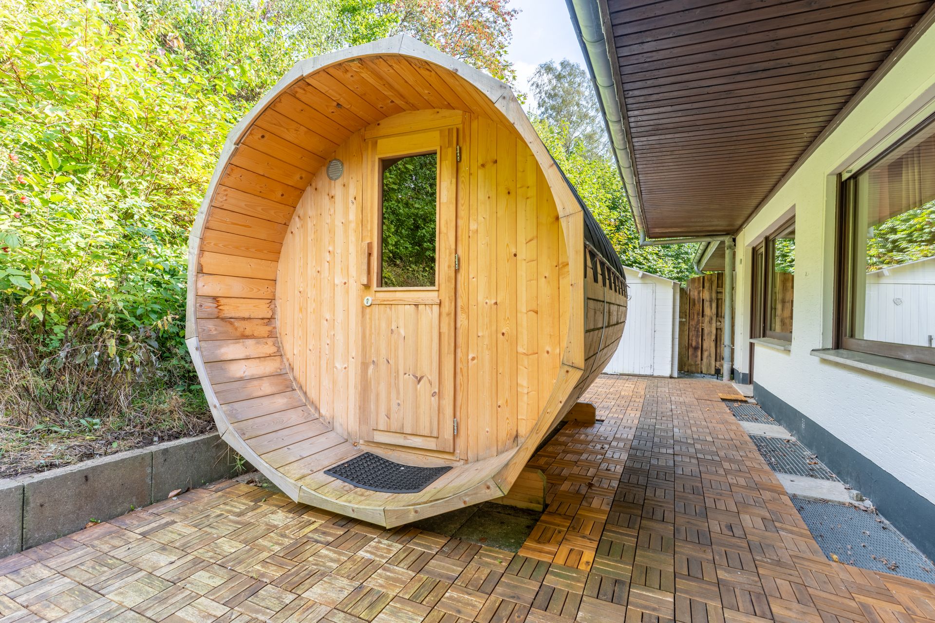 Große, holzfassförmige Sauna steht auf einer gefliesten Terrasse neben einem Wohnhaus.