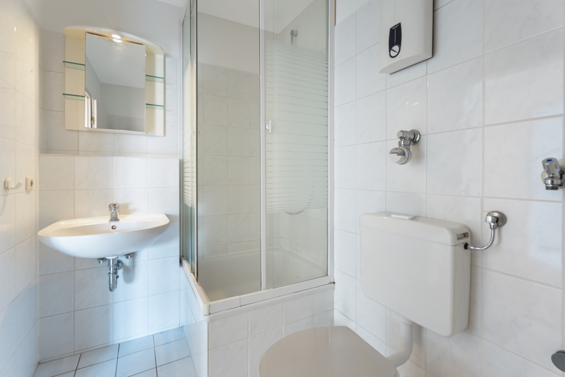 Helles Badezimmer mit Waschbecken, Spiegel, Dusche und WC, alles mit weißen Fliesen.