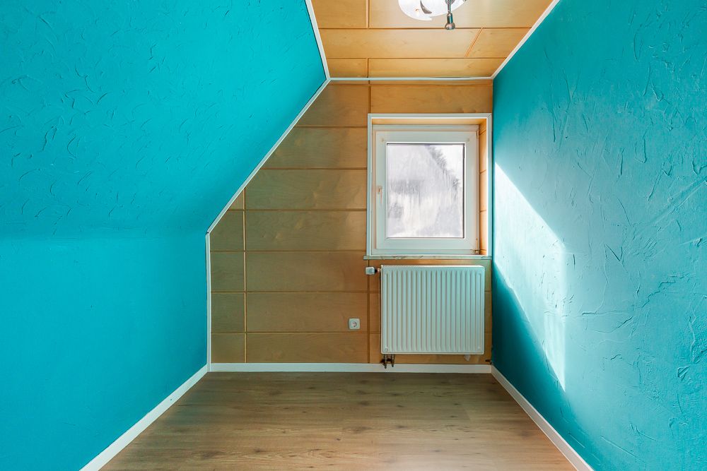 Kleiner Raum mit türkisfarbenen Wänden, Fenster und Heizkörper unter schräger Decke.