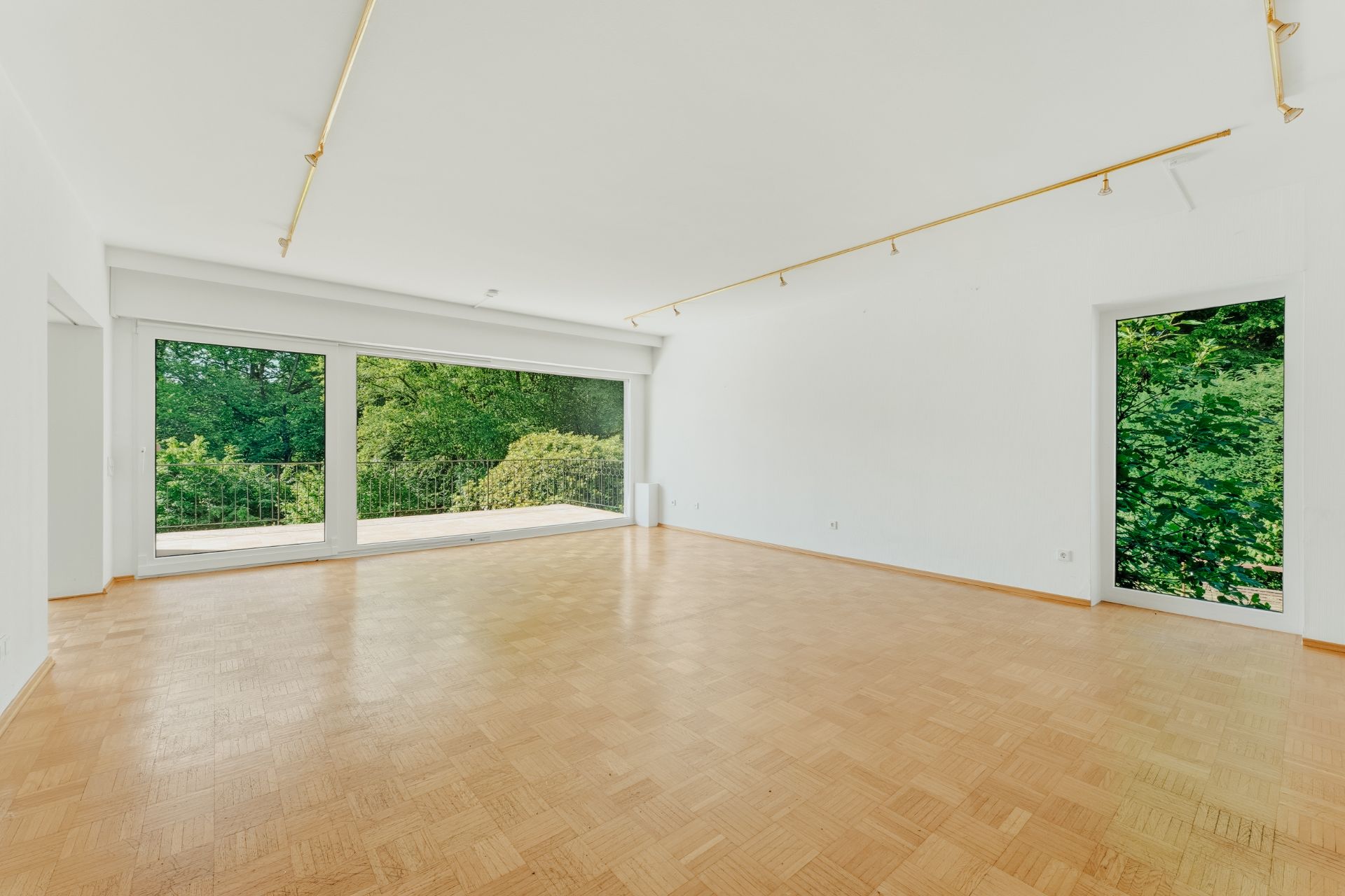 Leerer Raum mit großem Fenster, Parkettboden und Blick auf grüne Bäume.