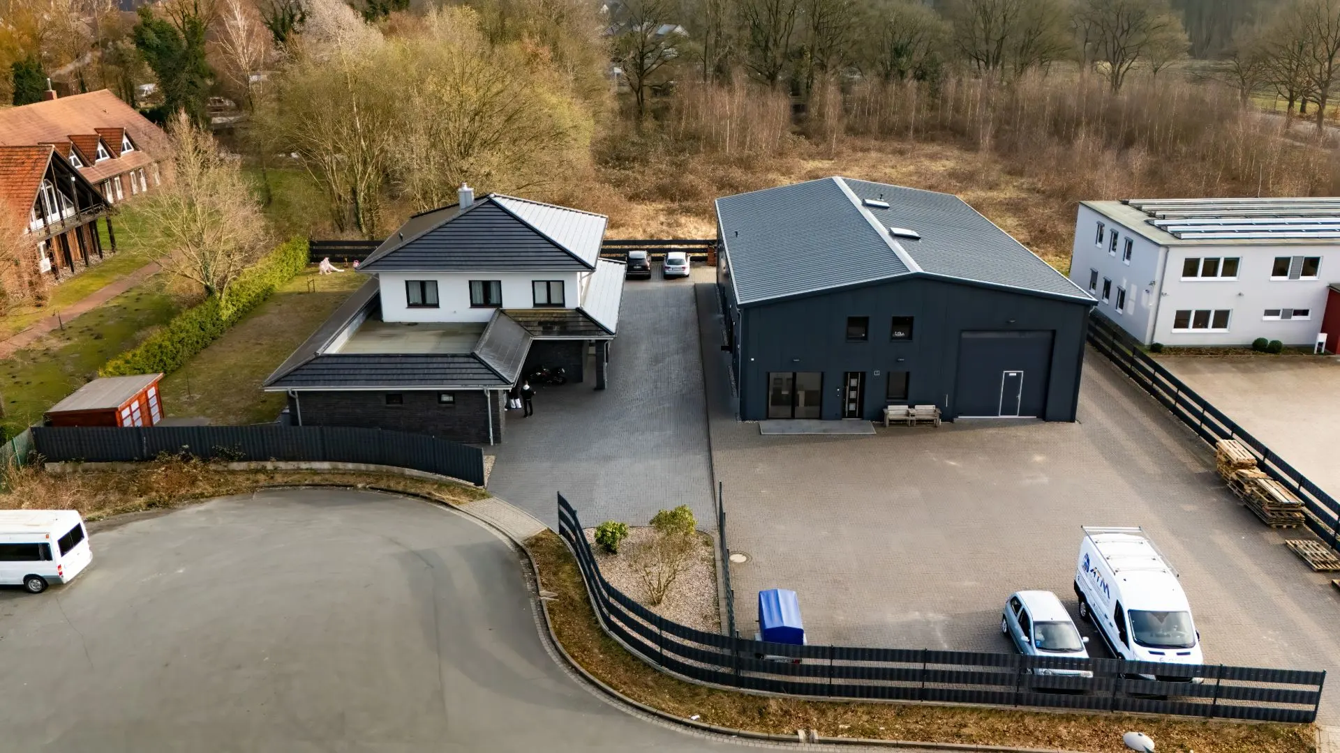 Luftaufnahme eines Wohnhauses mit Carport und angrenzender großer Halle, umgeben von Bäumen und Zaun.