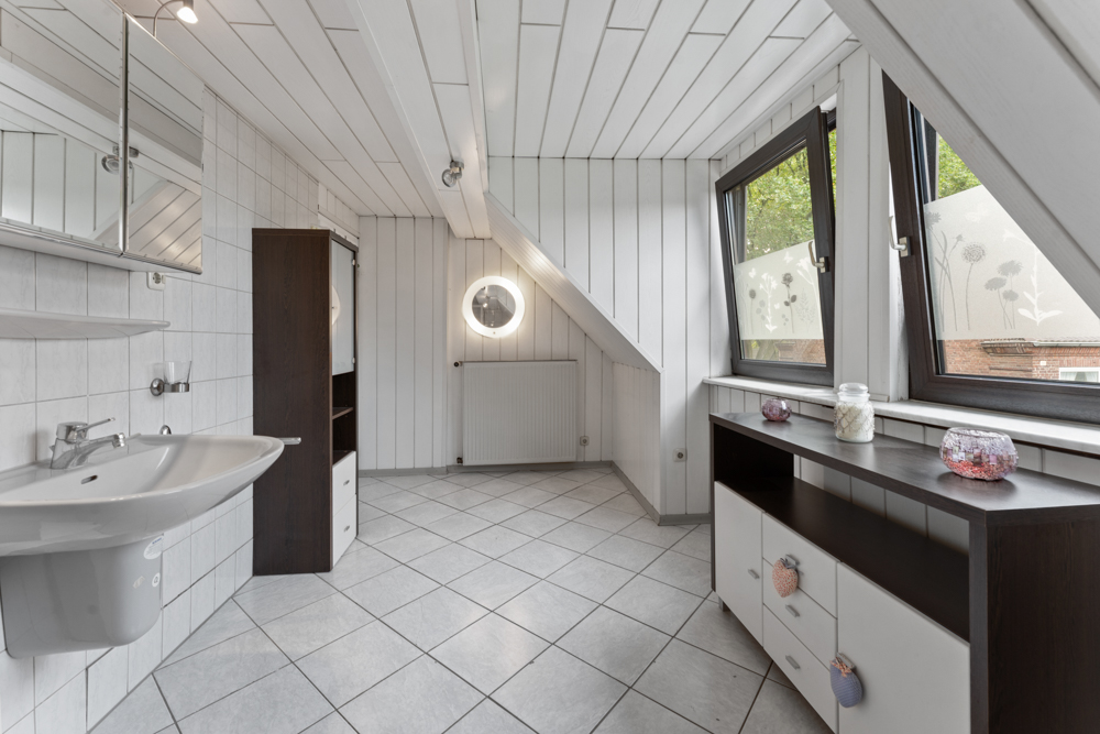 Helles Badezimmer mit Waschbecken, Spiegelschrank, Kommode und großen Fenstern.