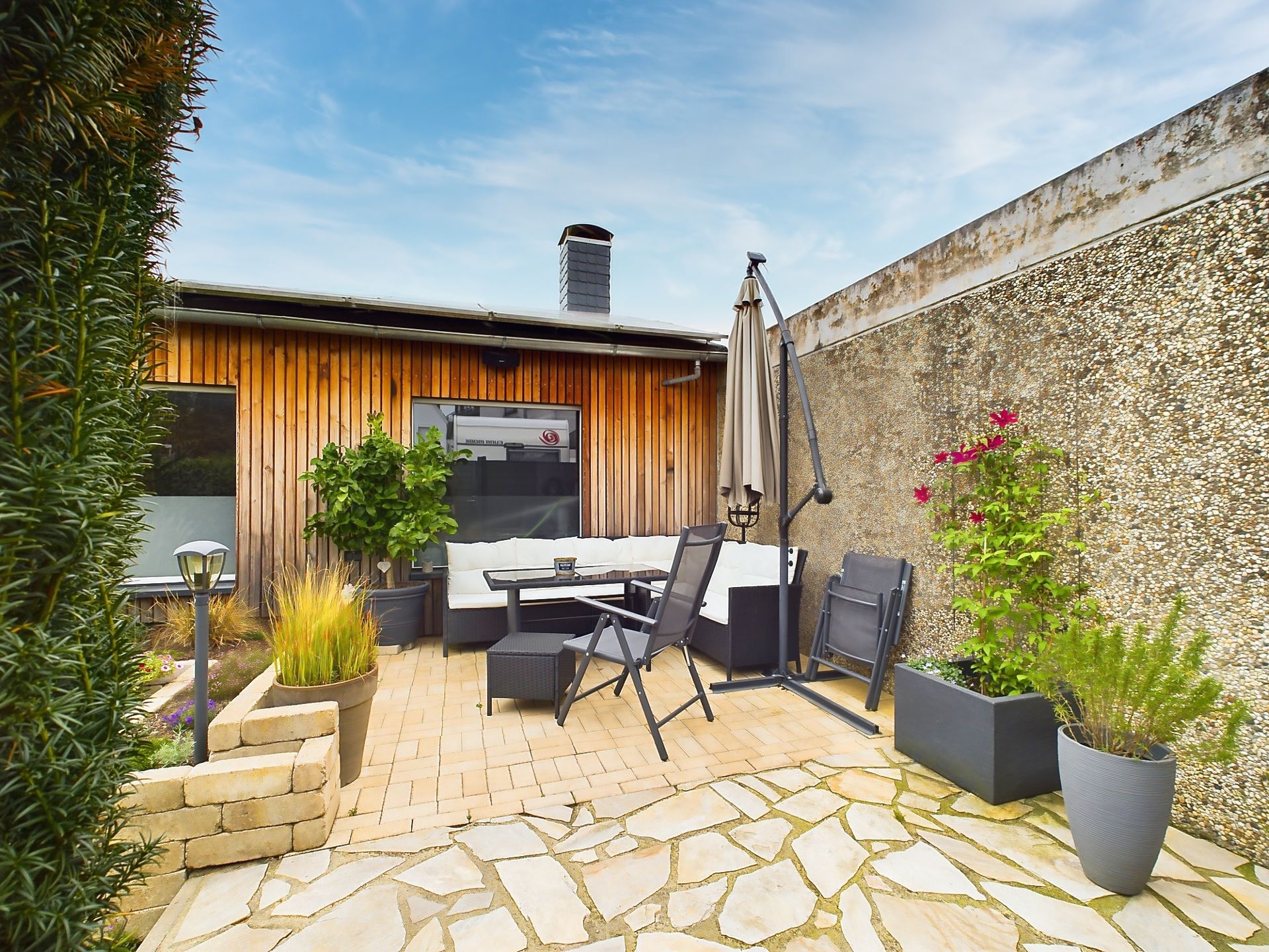 Moderne Terrasse mit Sitzgruppe, Sonnenschirm und vielen Pflanzen in Töpfen.