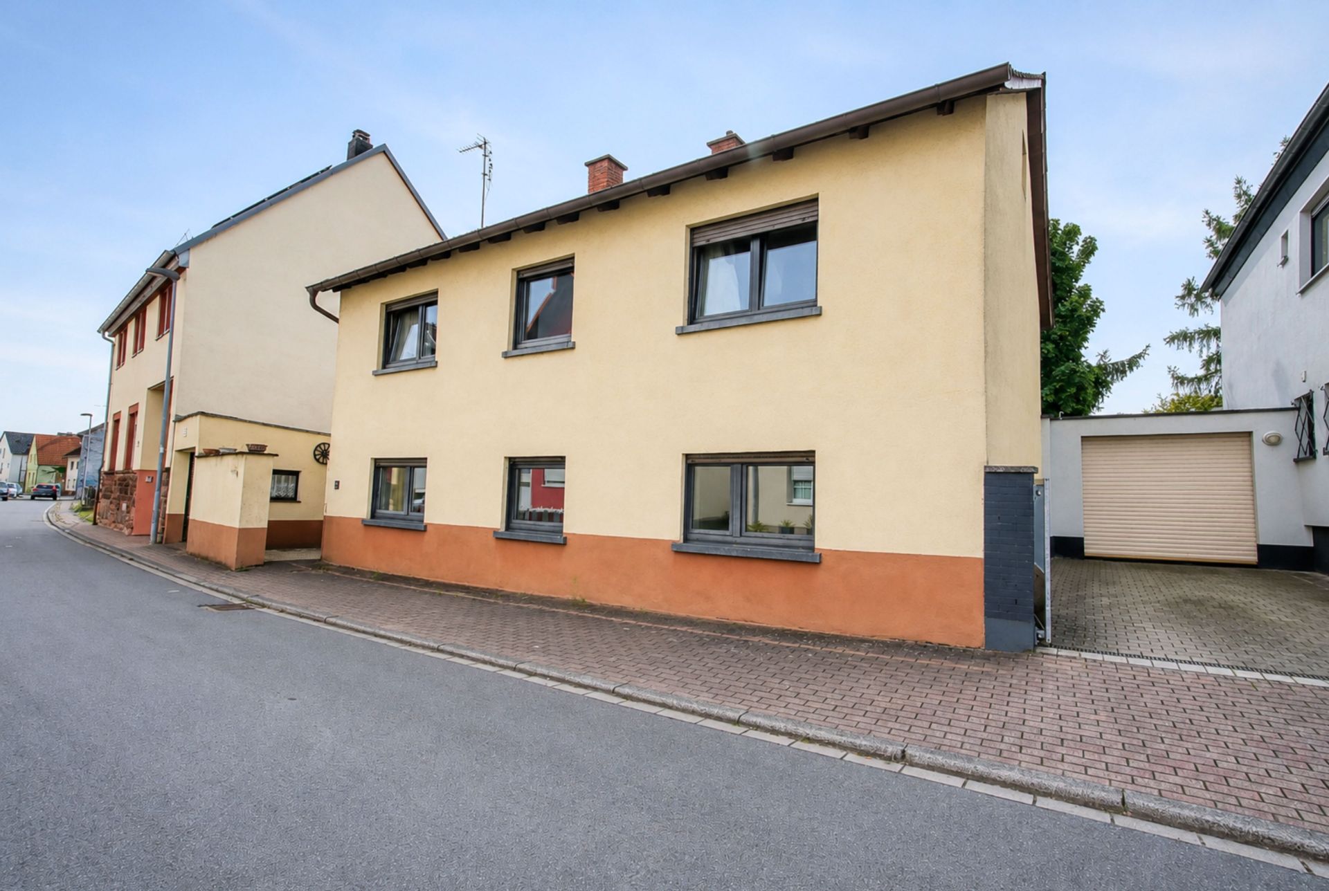 Zweistöckiges, gelb-beiges Wohnhaus mit braunem Sockel an einer ruhigen Straße.