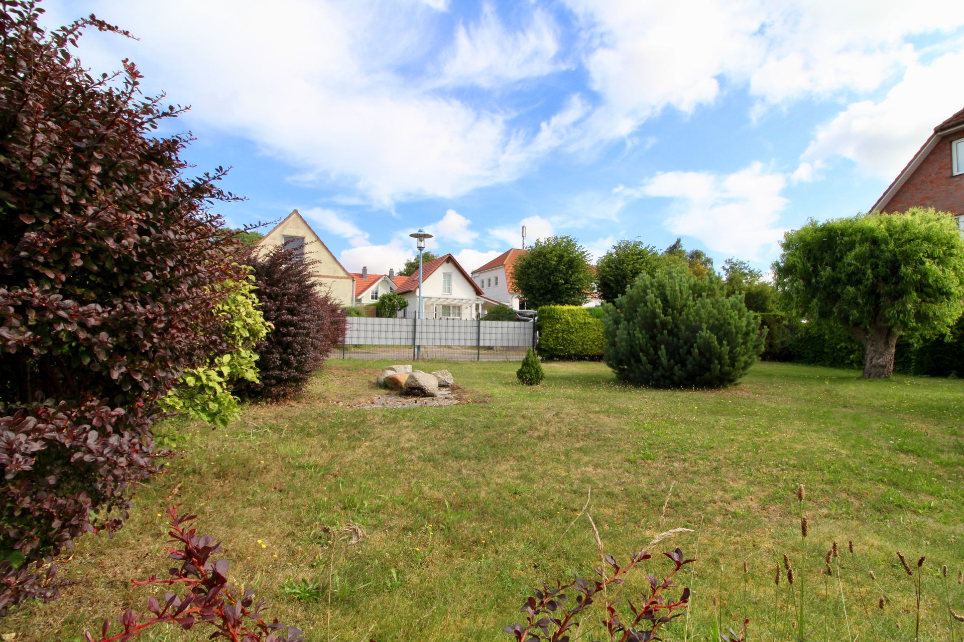 Garten mit Rasen, Sträuchern und Bäumen, im Hintergrund Häuser und blauer Himmel.