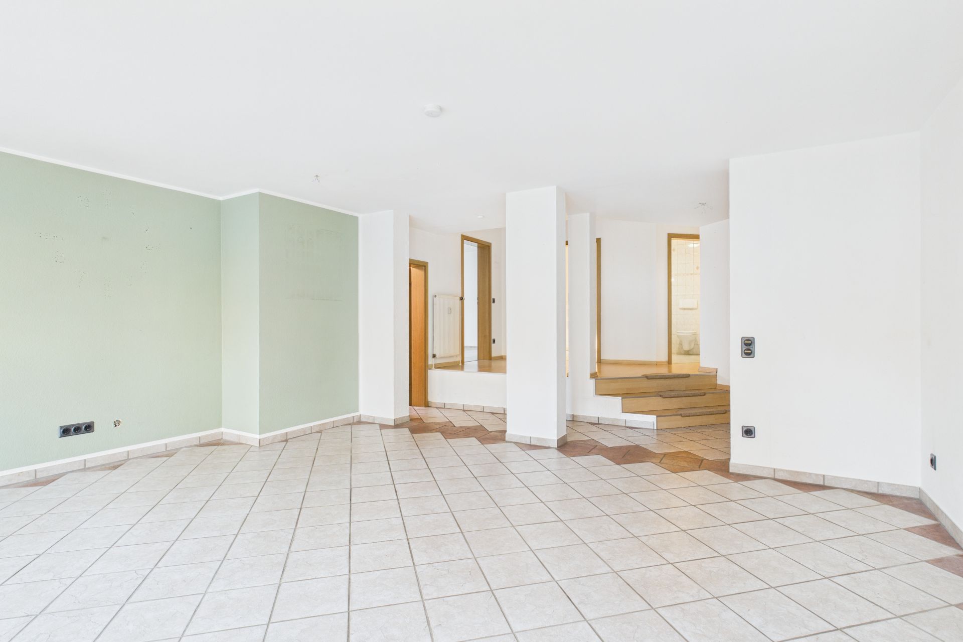 Helles, leeres Wohnzimmer mit Fliesenboden und Treppe im Hintergrund.