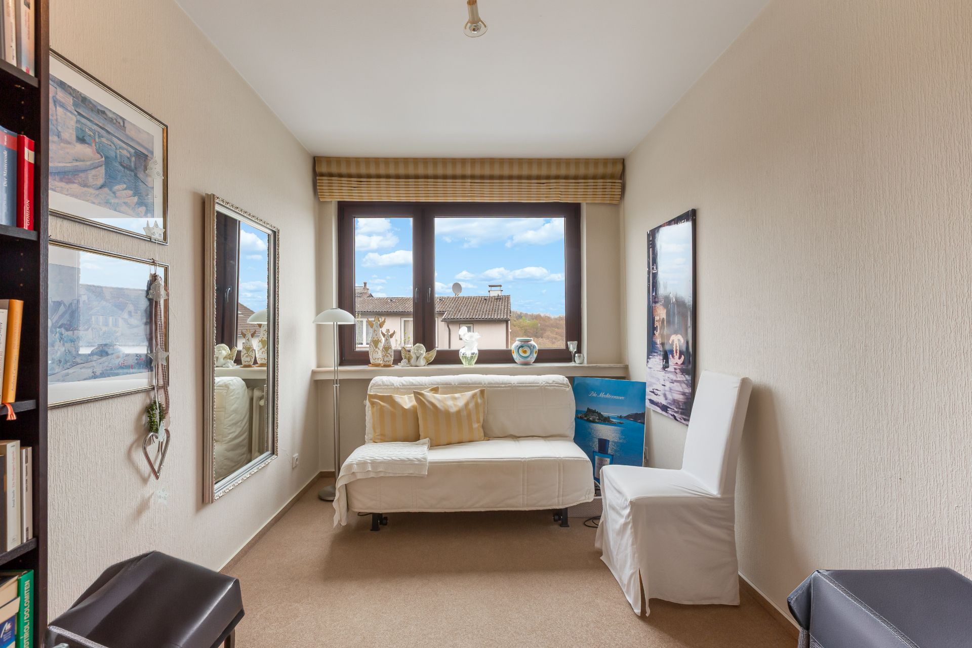 Helles Zimmer mit Sofa, Stuhl, großem Fenster und Büchernregal an der Seite.