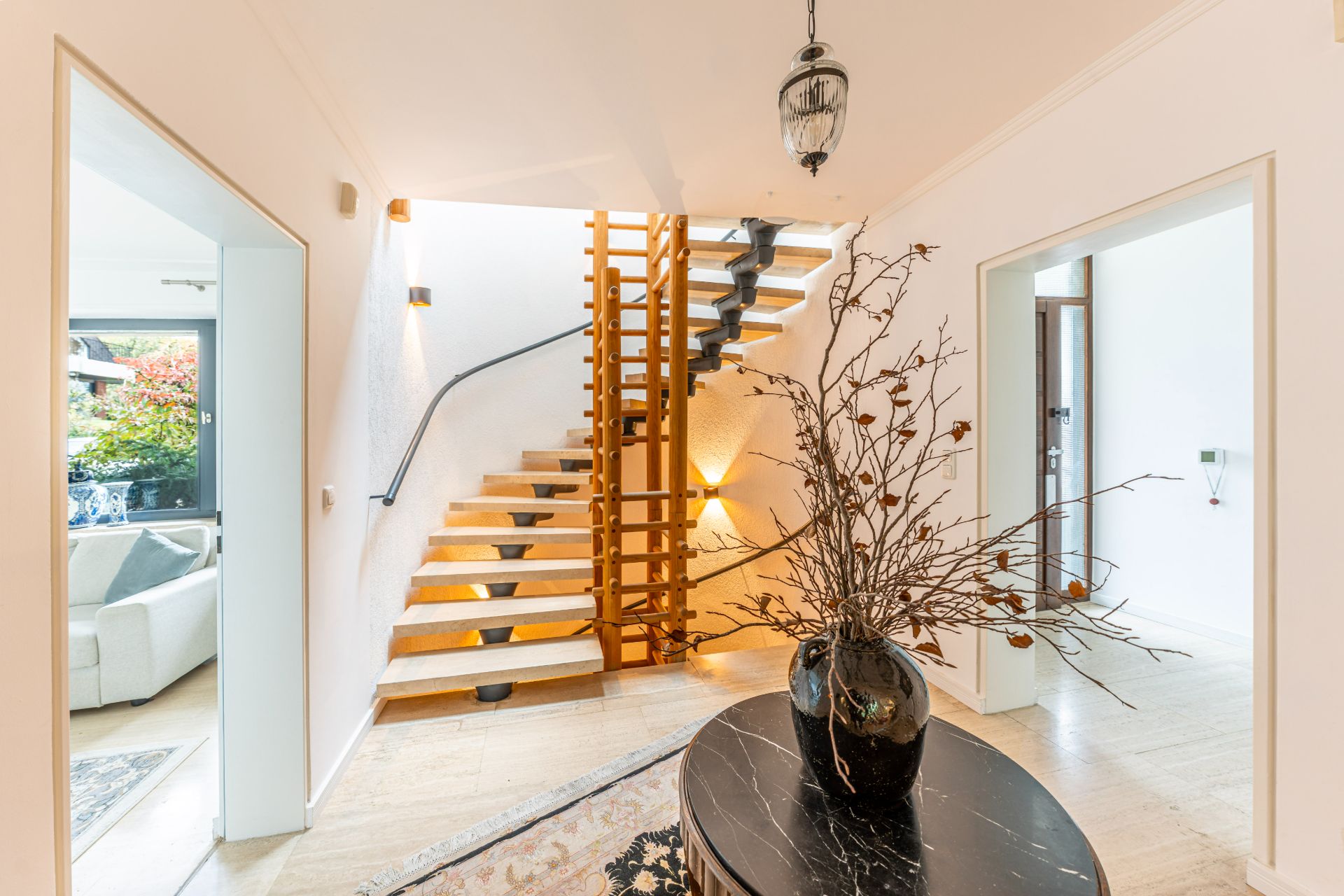 Heller Flur mit offener Holztreppe, schwarzem Tisch und Vase mit Zweigen.
