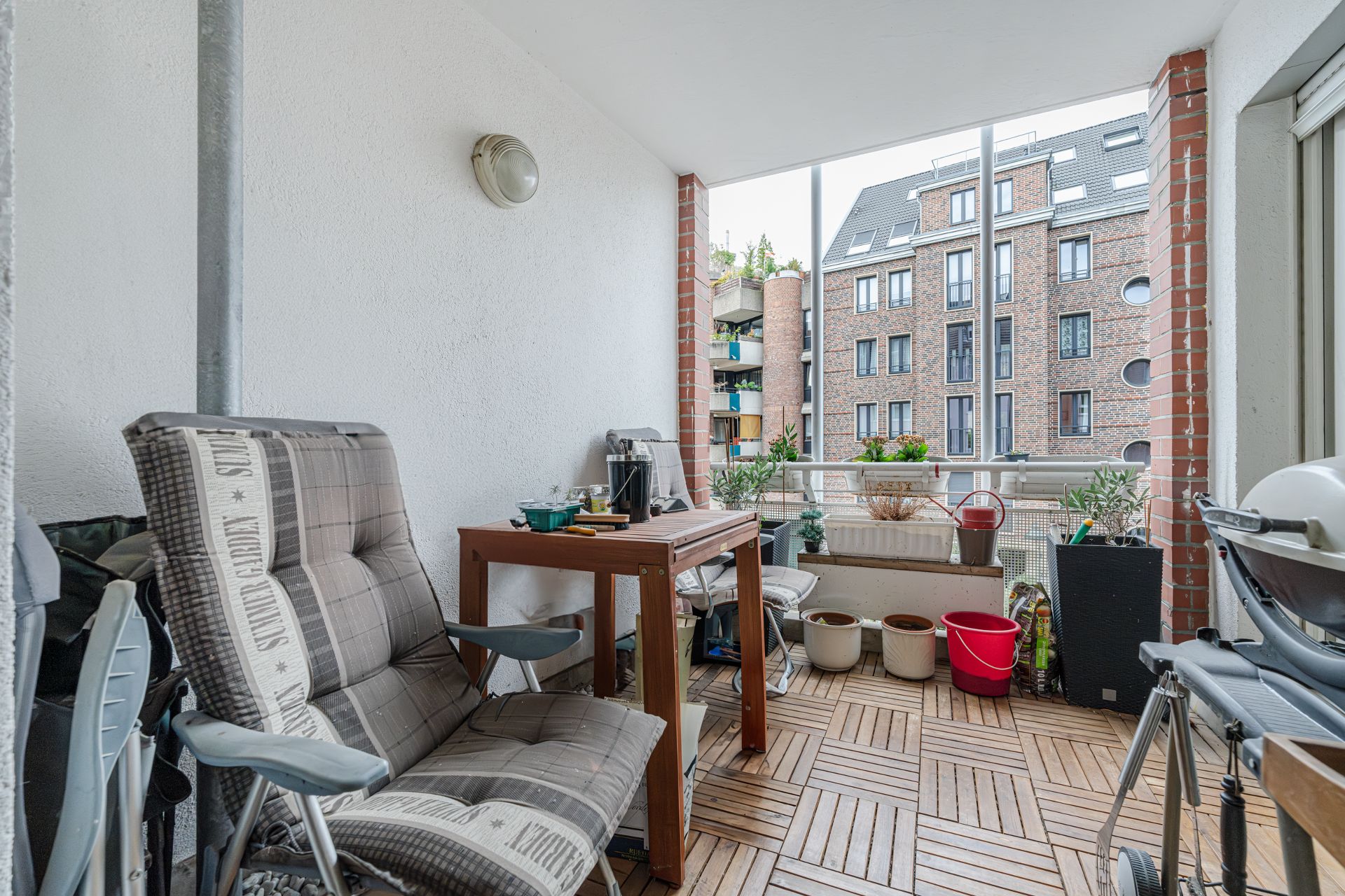Balkon mit Holzboden, Stühlen, Tisch, Pflanzgefäßen und Blick auf Wohngebäude.