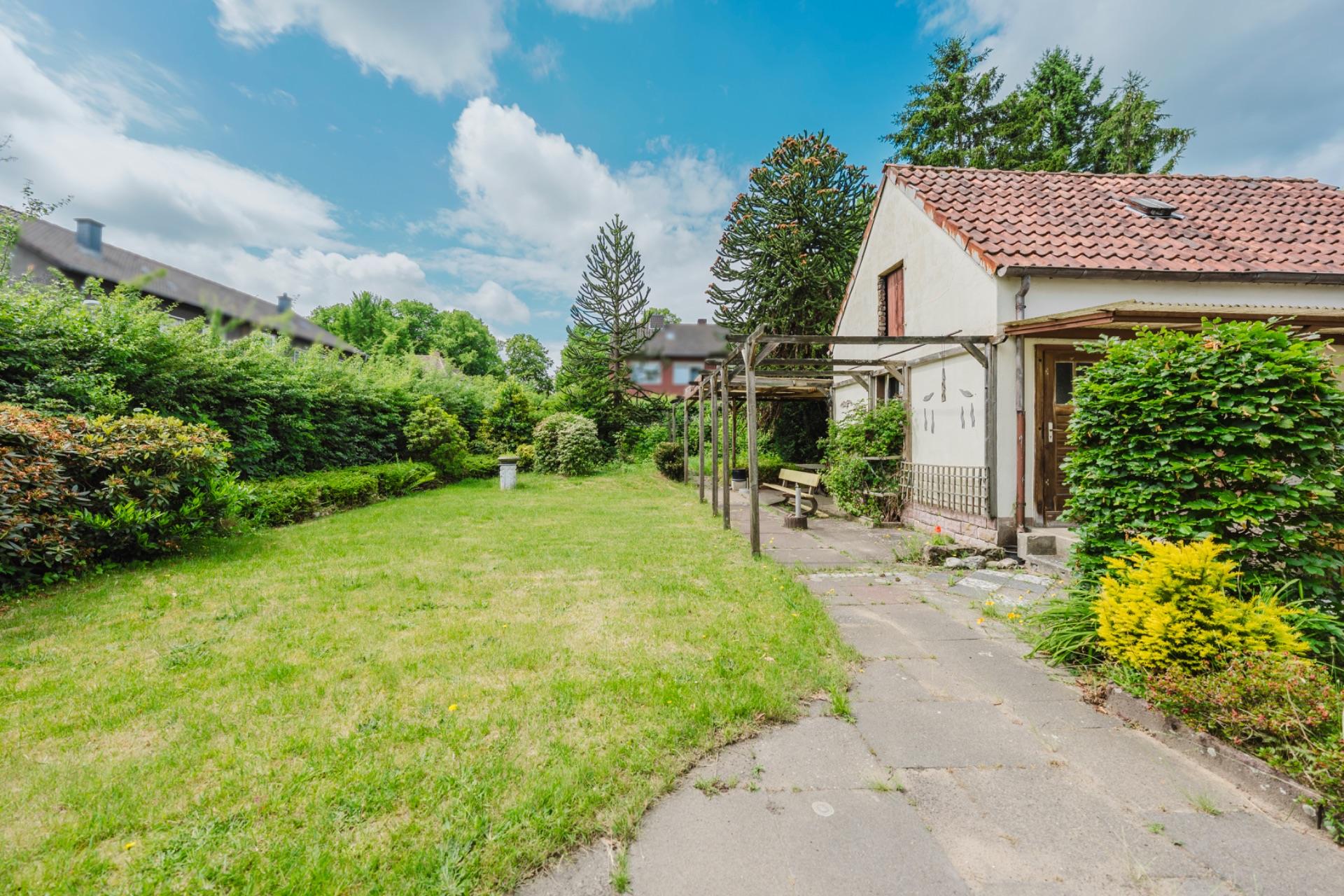 Garten mit Rasenfläche, Büschen und einem Haus mit rotem Dach auf der rechten Seite.