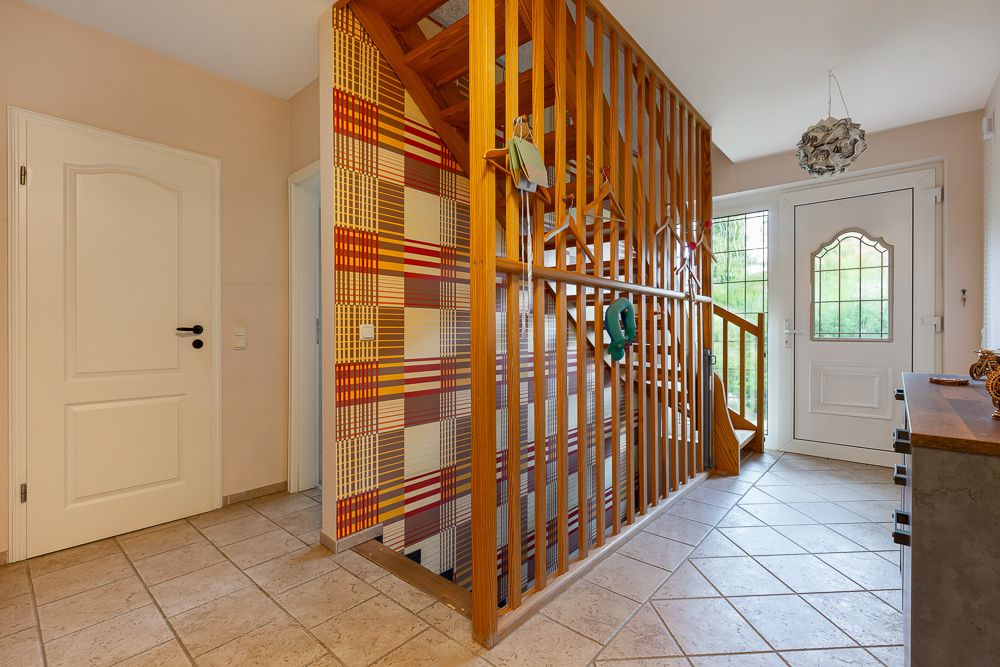 Flur mit Holztreppe, Fliesenboden und weißer Eingangstür, bunte Wand hinter der Treppe.