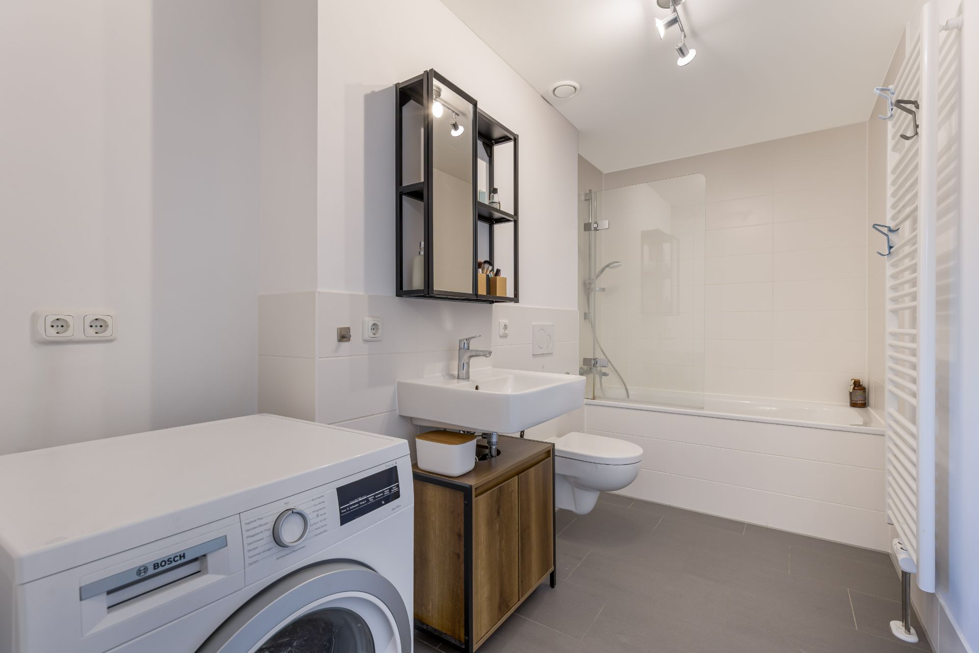 Modernes Badezimmer mit Waschmaschine, Waschbecken, Spiegel, Toilette und Badewanne mit Dusche.