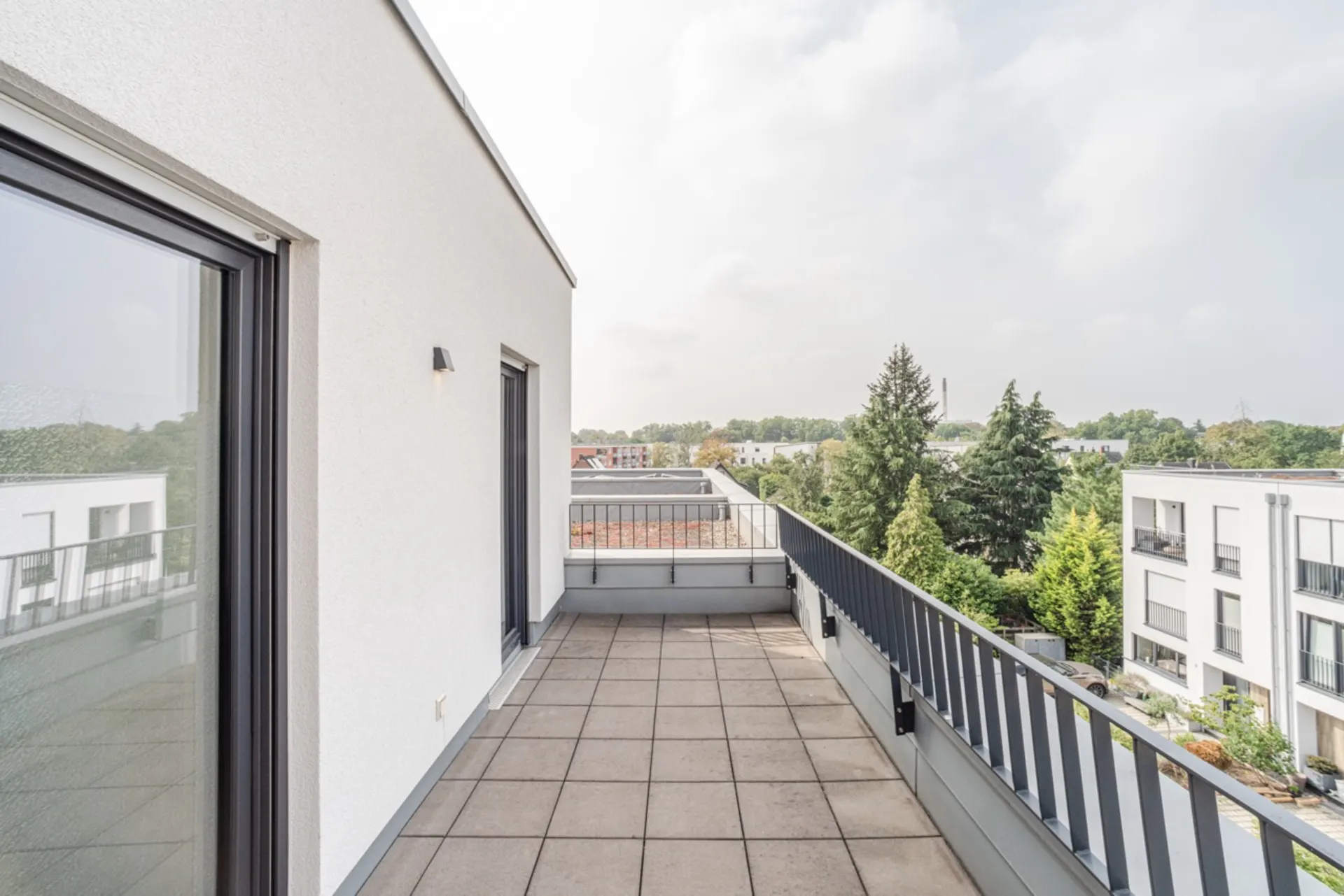 Große Dachterrasse mit Fliesenboden und Metallgeländer, Blick auf Bäume und Nachbargebäude.