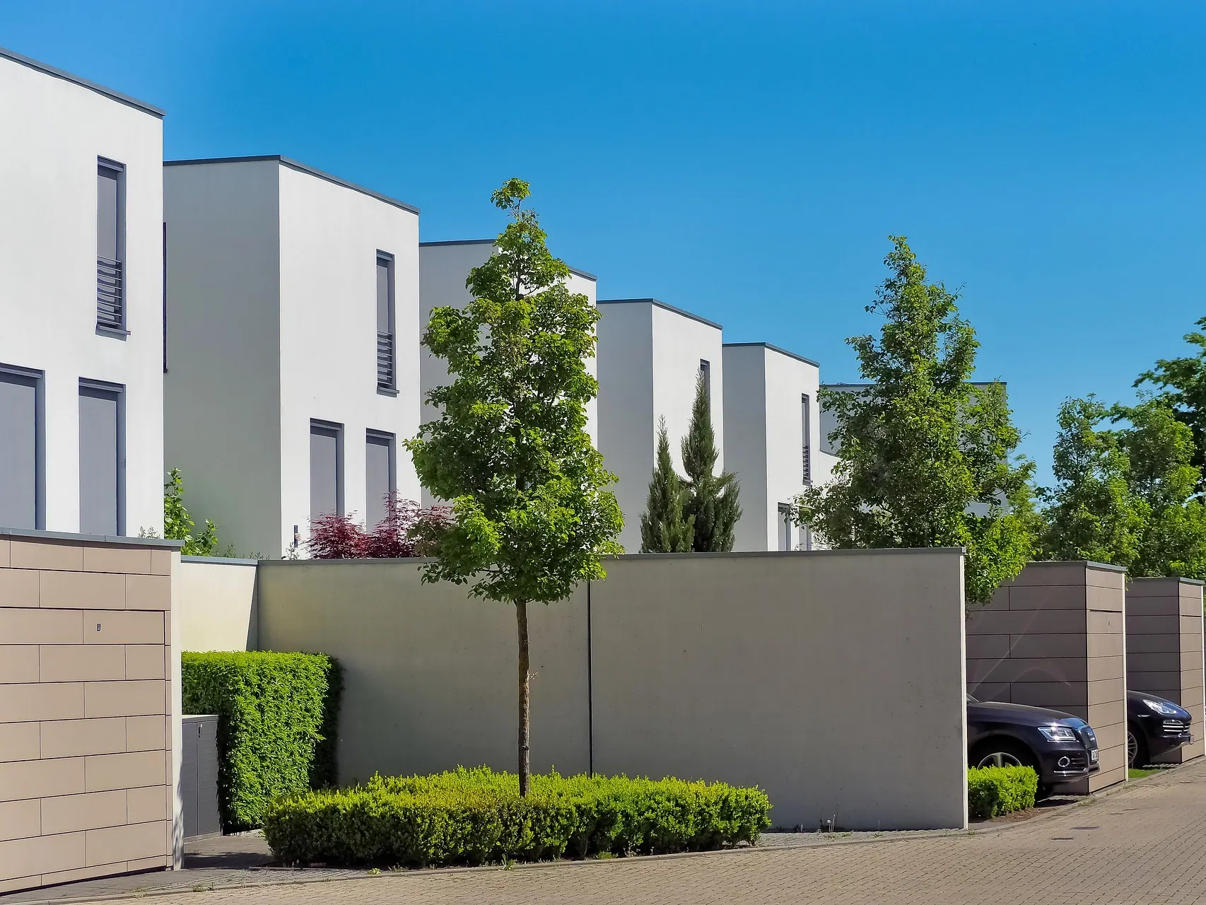 Moderne weiße Wohnhäuser mit Flachdach, Bäumen und parkenden Autos vor einer Mauer.