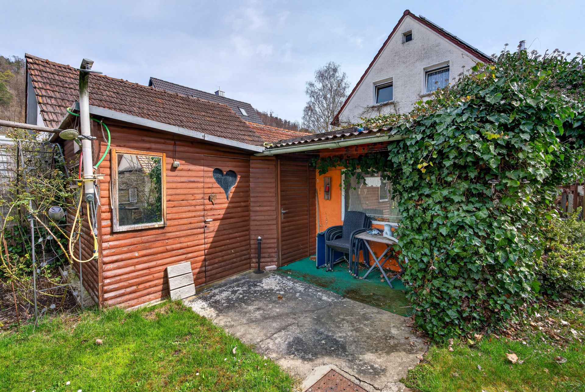 Kleines Gartenhaus mit Holzfassade, überdachter Terrasse und Efeu, davor Stühle und Tisch.