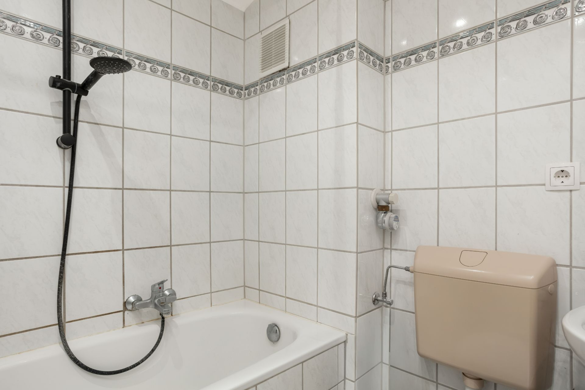 Teil eines Badezimmers mit Badewanne, Duschkopf und Toilettenspülkasten vor weißen Fliesen.