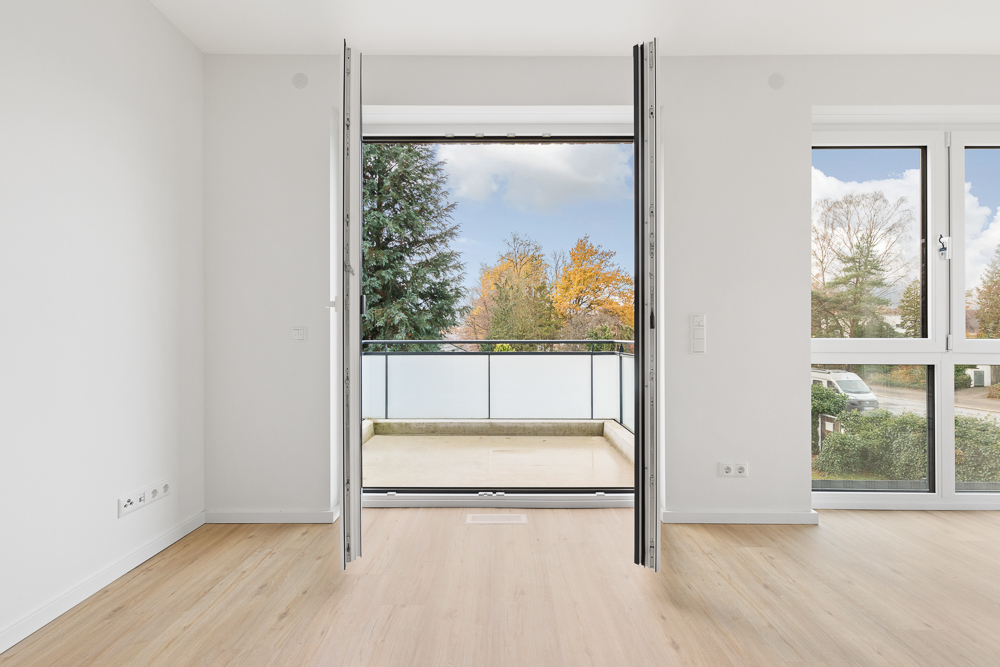 Helles Zimmer mit offenem Balkon und großen Fenstern, Blick auf Bäume im Garten.
