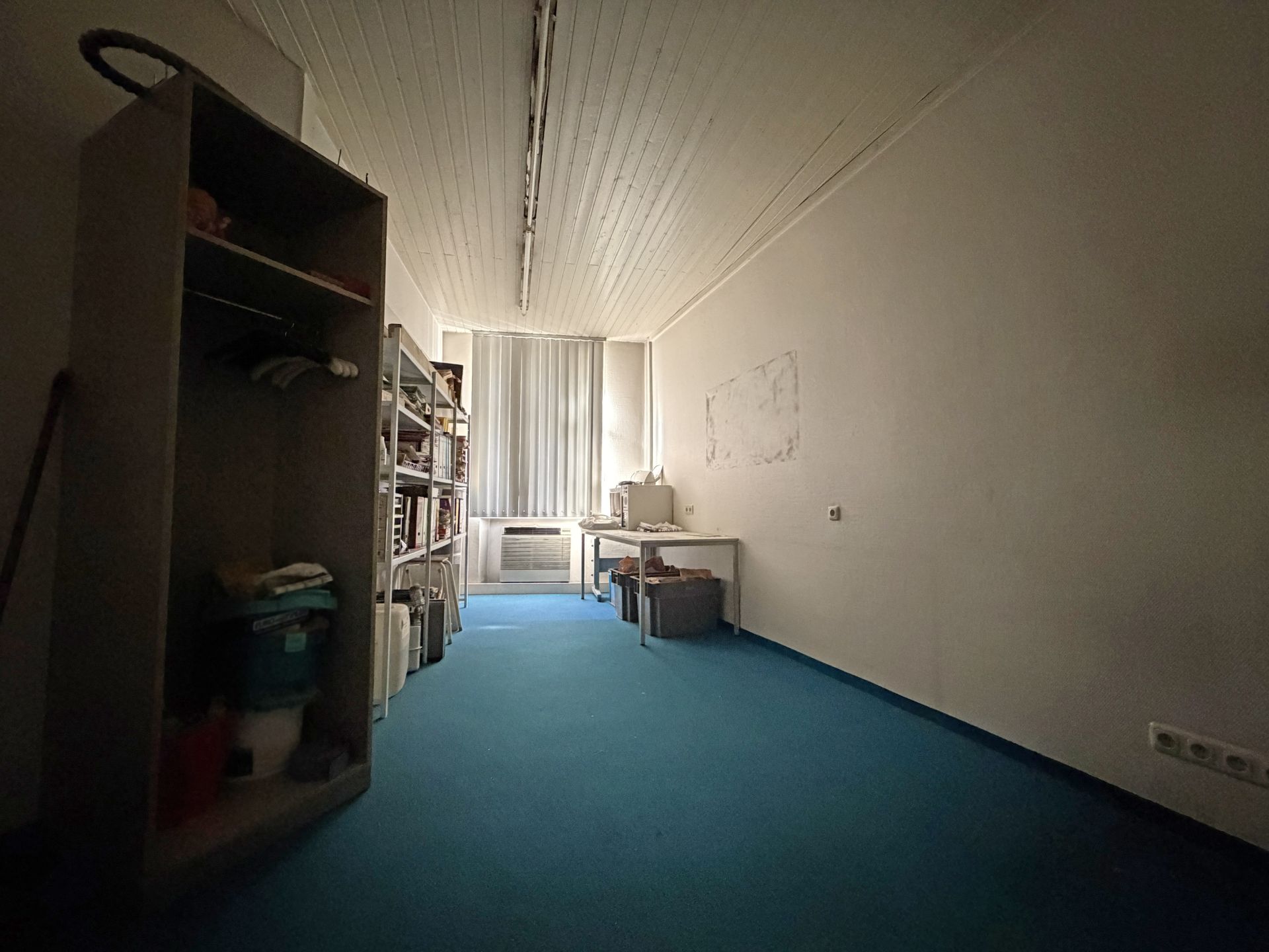 Raum mit blauem Teppichboden, Regalen links und Schreibtisch vor Fenster mit Jalousien.