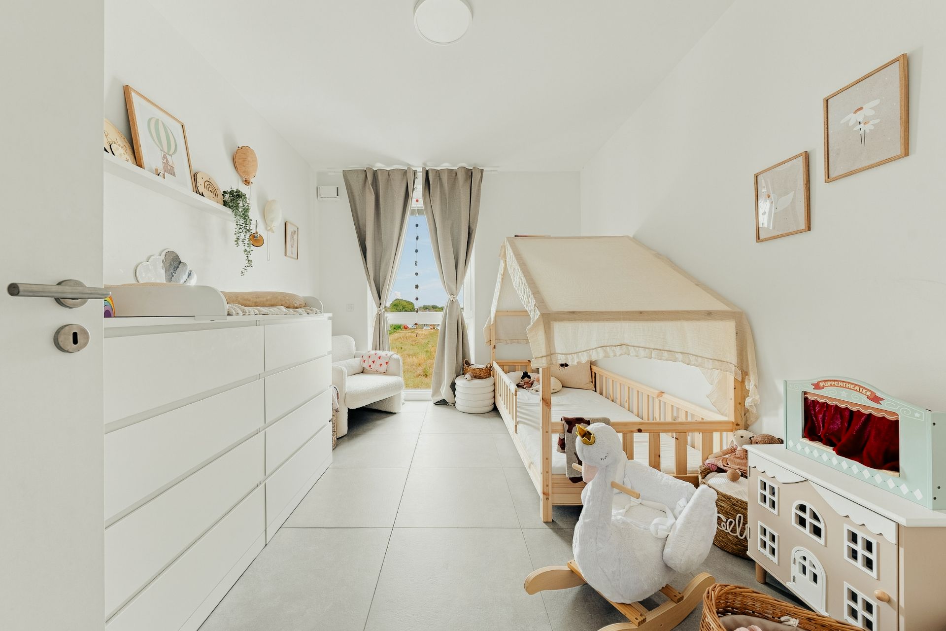 Helles Kinderzimmer mit Holzbett, Wickelkommode, Spielzeug und Fenster mit Vorhängen.
