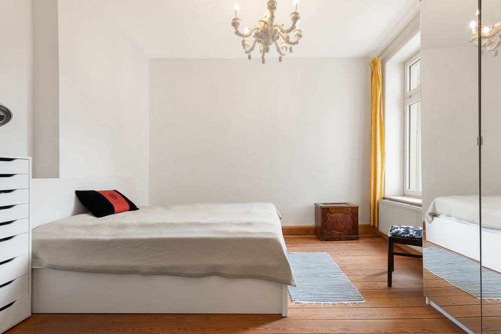 Helles Schlafzimmer mit Bett, Kronleuchter, Spiegel, gelben Vorhängen und Holzboden.