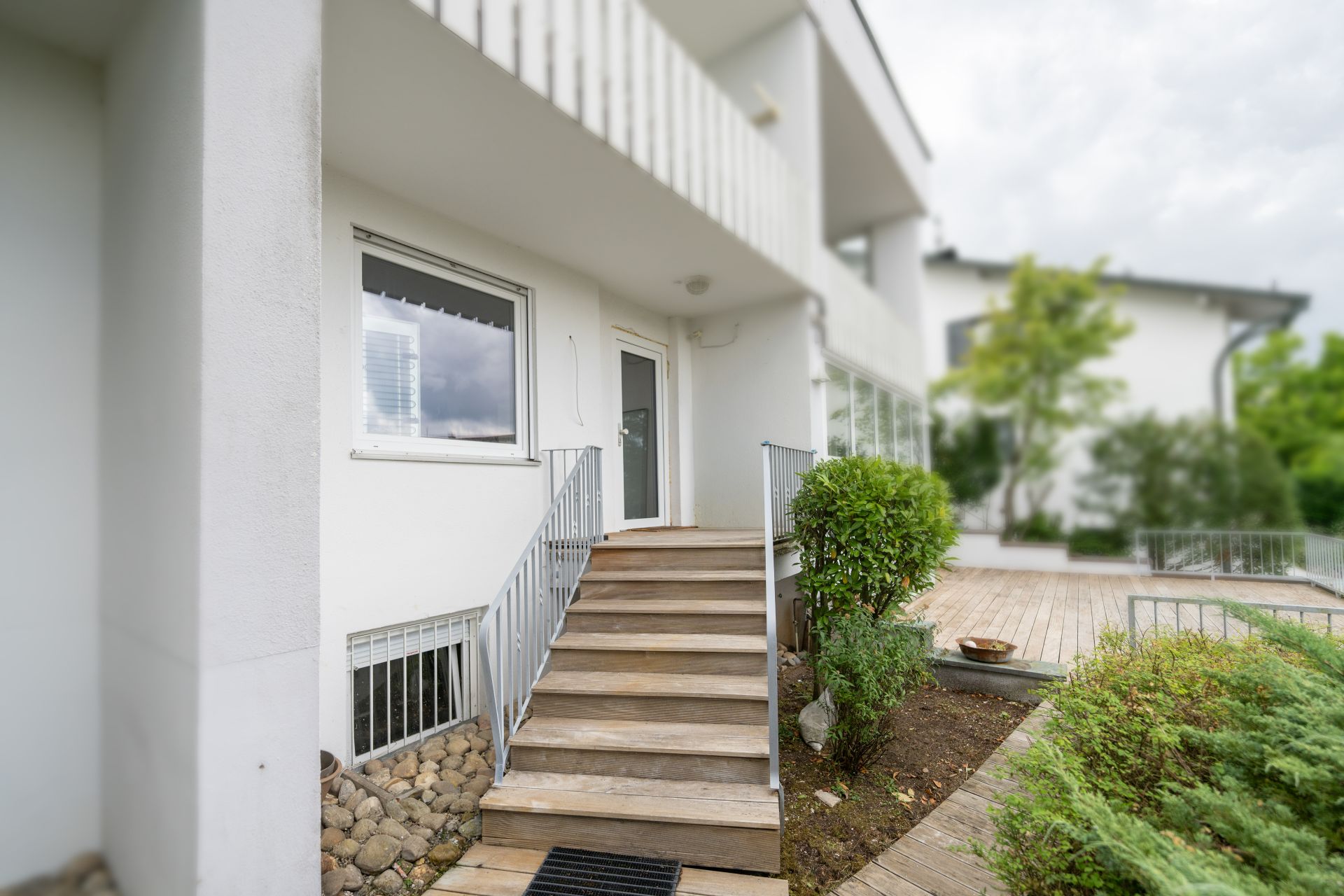 Holztreppe mit Geländer führt zum Eingang eines weißen Hauses mit Terrasse und Pflanzen.