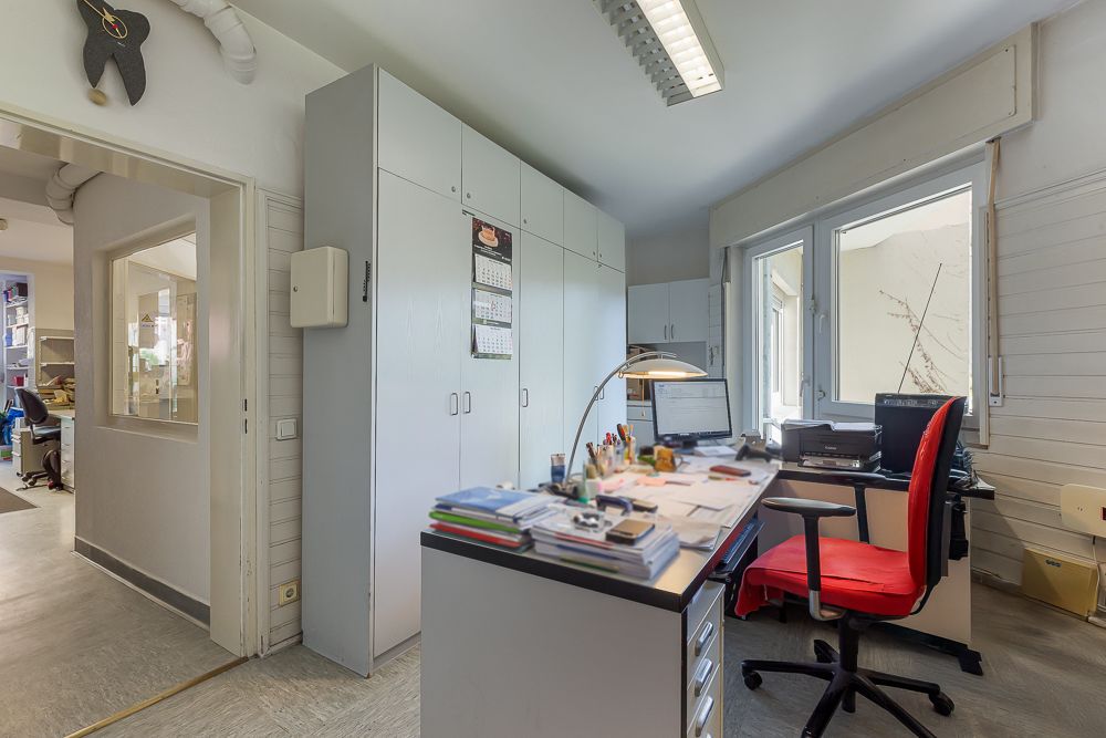 Helles Arbeitszimmer mit Schreibtisch, rotem Stuhl, Regalen und großem Fenster.