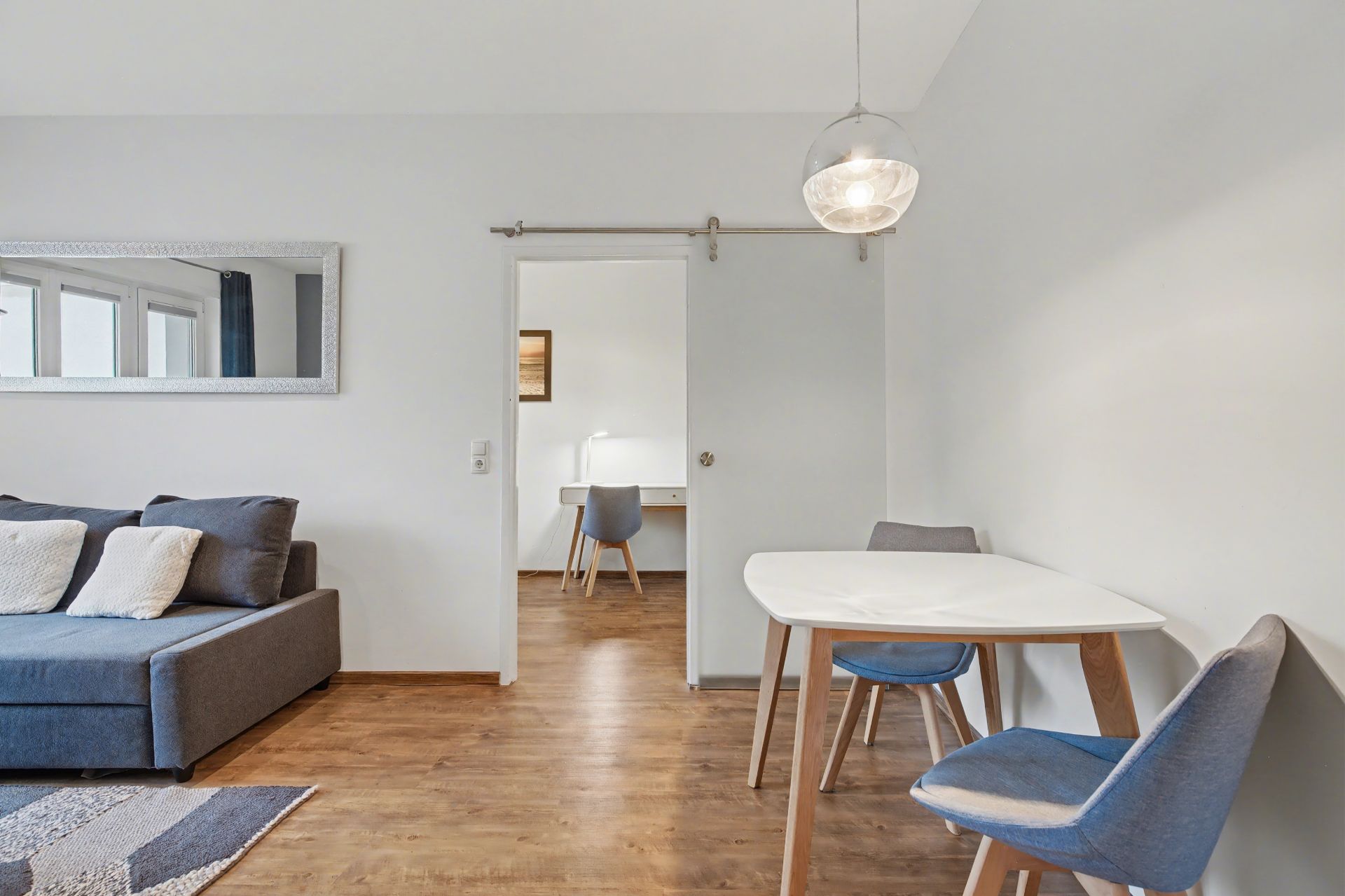 Helles Wohnzimmer mit Sofa, Esstisch, drei Stühlen und Blick auf einen kleinen Arbeitsbereich.