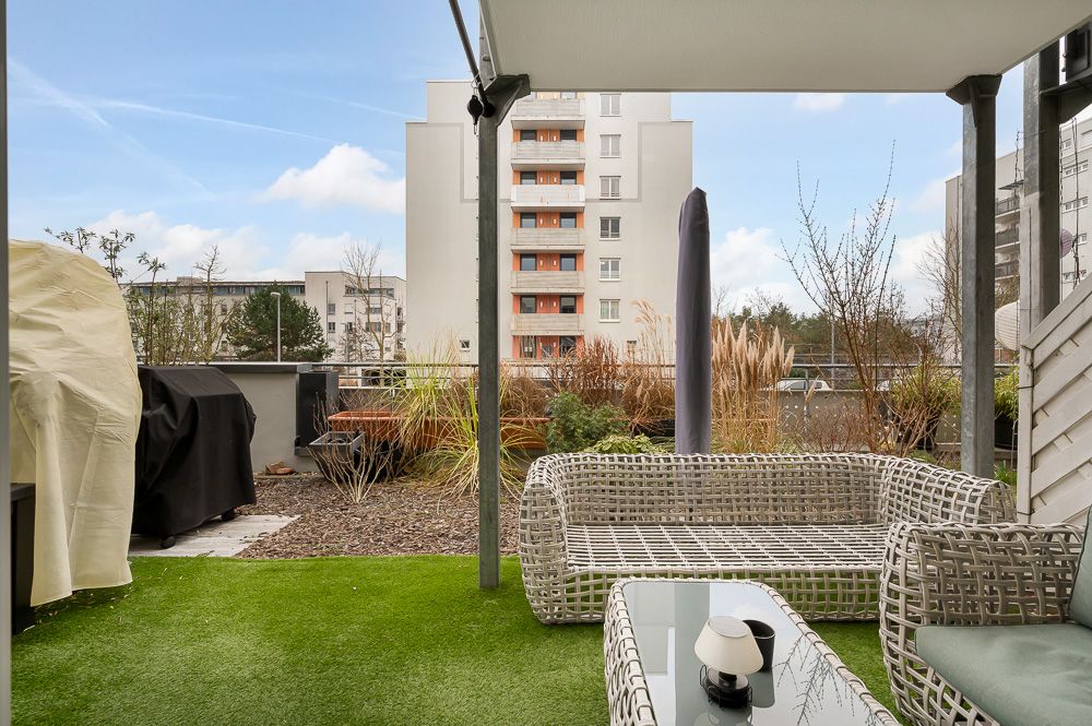 Terrasse mit Kunstrasen, Rattanmöbeln und Blick auf ein Wohnhaus.