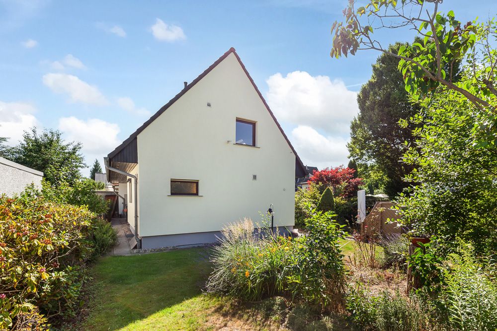 Haus mit Satteldach und Garten, umgeben von Büschen und Pflanzen an einem sonnigen Tag.