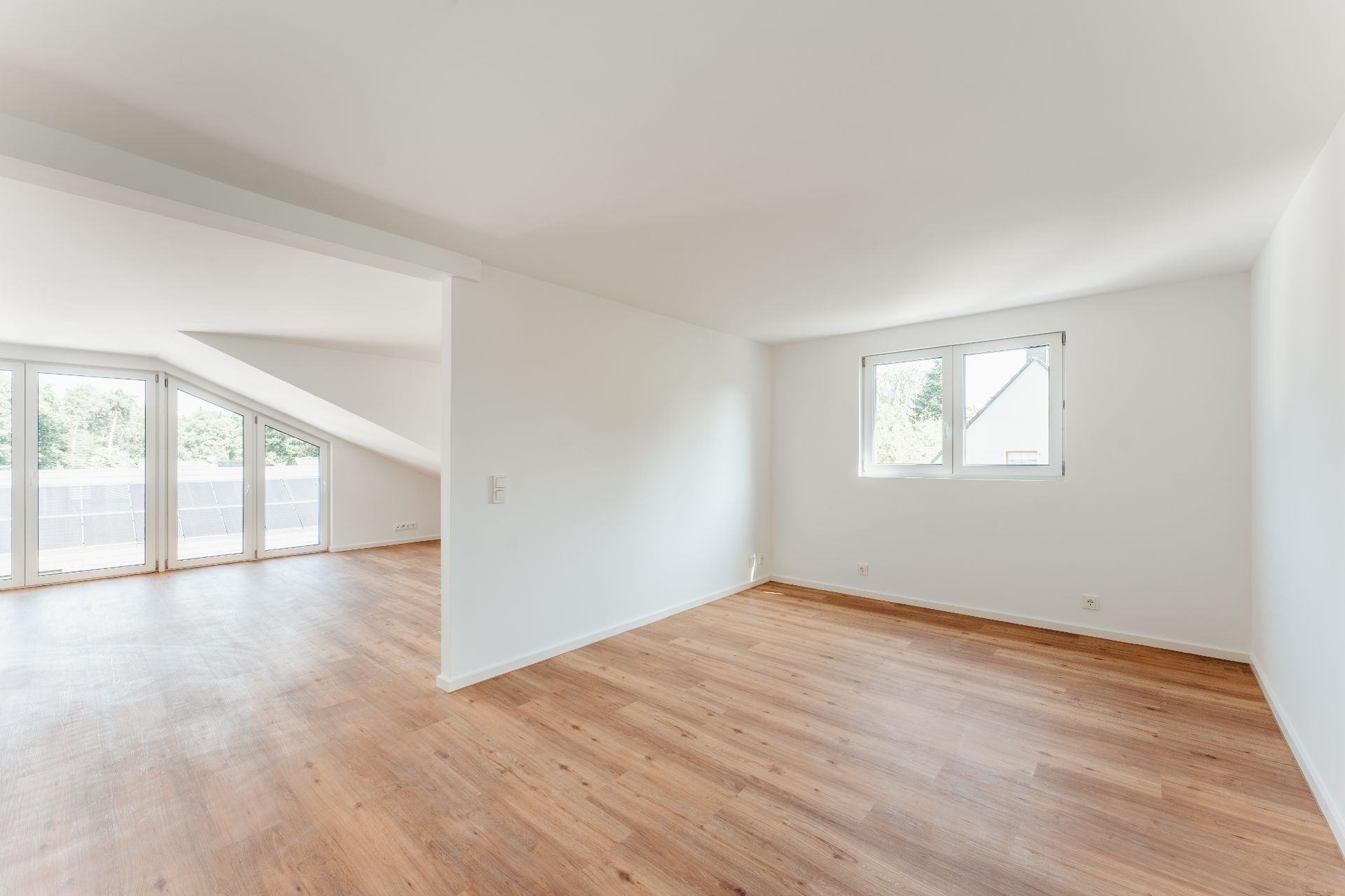 Helles, leeres Zimmer mit Holzfußboden, großen Fenstern und weiß gestrichenen Wänden.