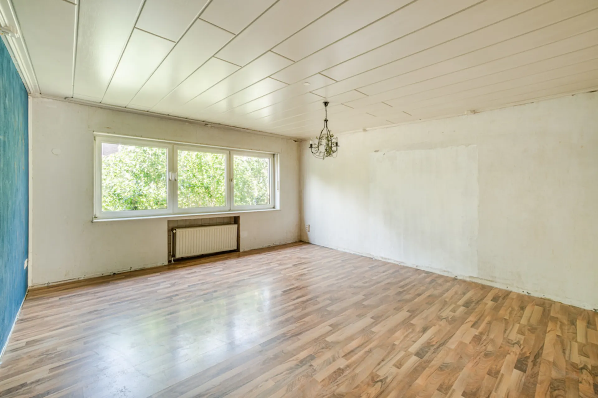 Leeres Zimmer mit Holzfußboden, großem Fenster, Heizung und Deckenleuchte.