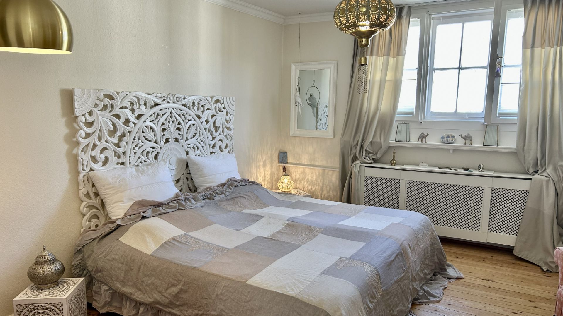 Schlafzimmer mit Doppelbett, geschnitztem Kopfteil, Holzboden und großem Fenster mit Vorhängen.