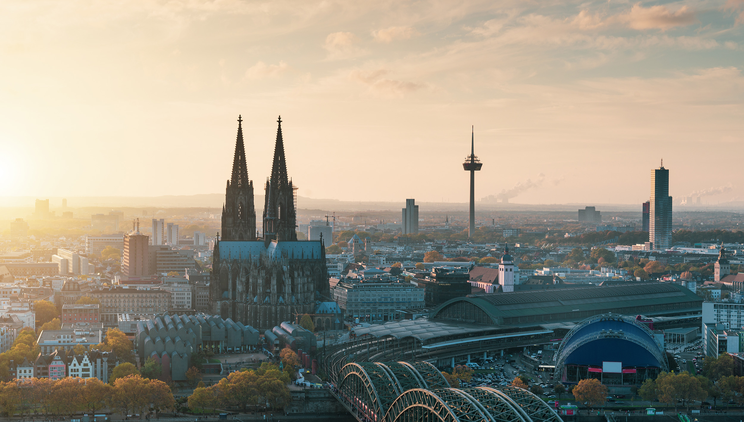 Stadtpanorama mit Kirche, Fernsehturm, Bahnhof und Brücke im Vordergrund bei Sonnenuntergang.