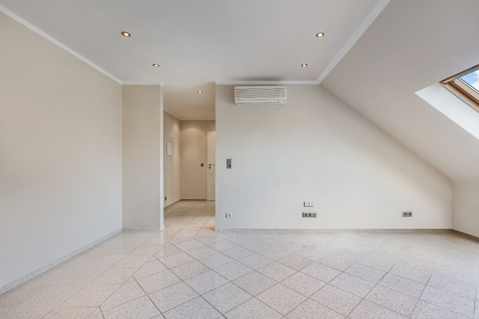 Helles, leeres Zimmer mit Fliesenboden, Dachschräge und Klimaanlage an der Wand.