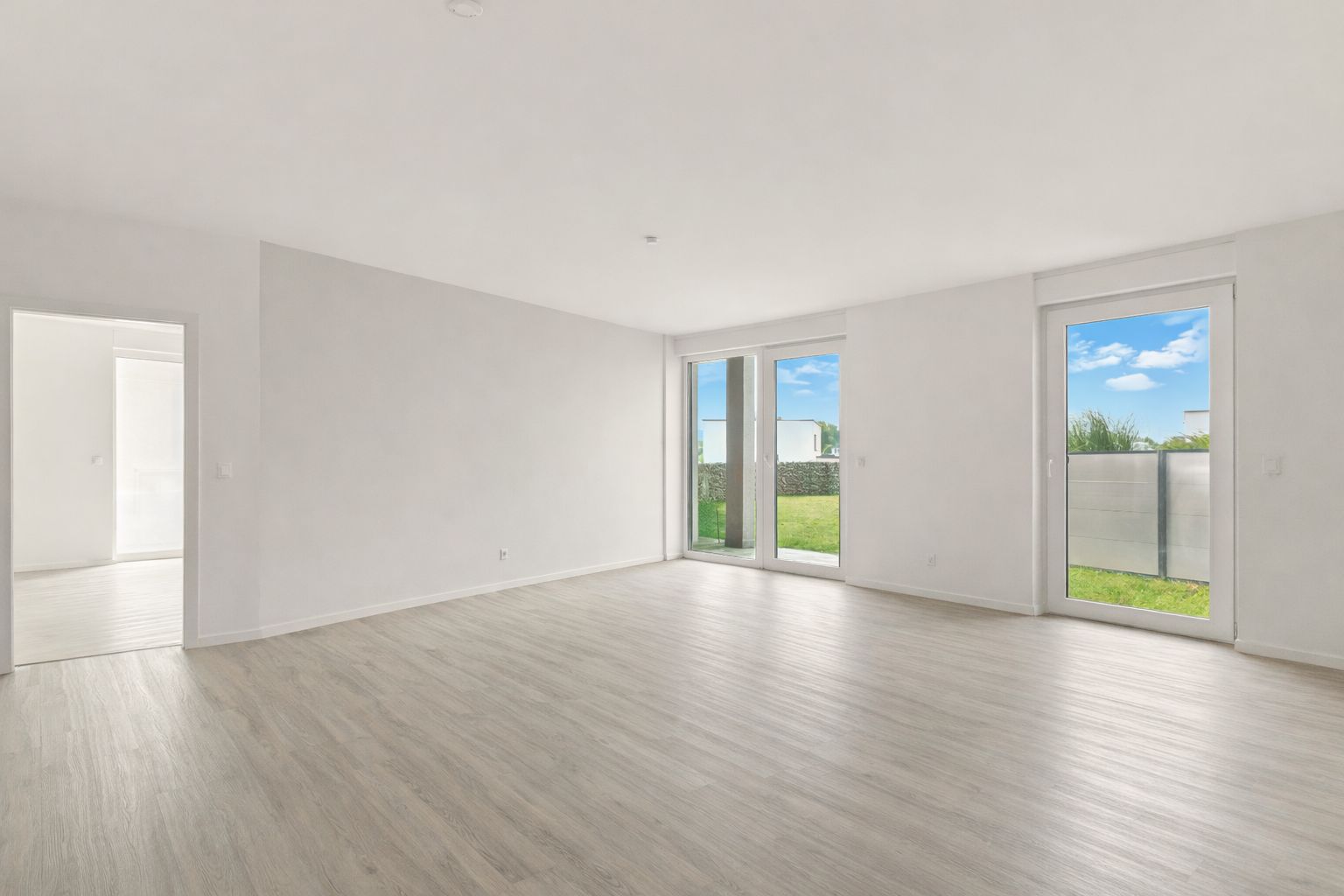 Heller leerer Raum mit großen Fenstern und Blick auf Garten, heller Holzboden.