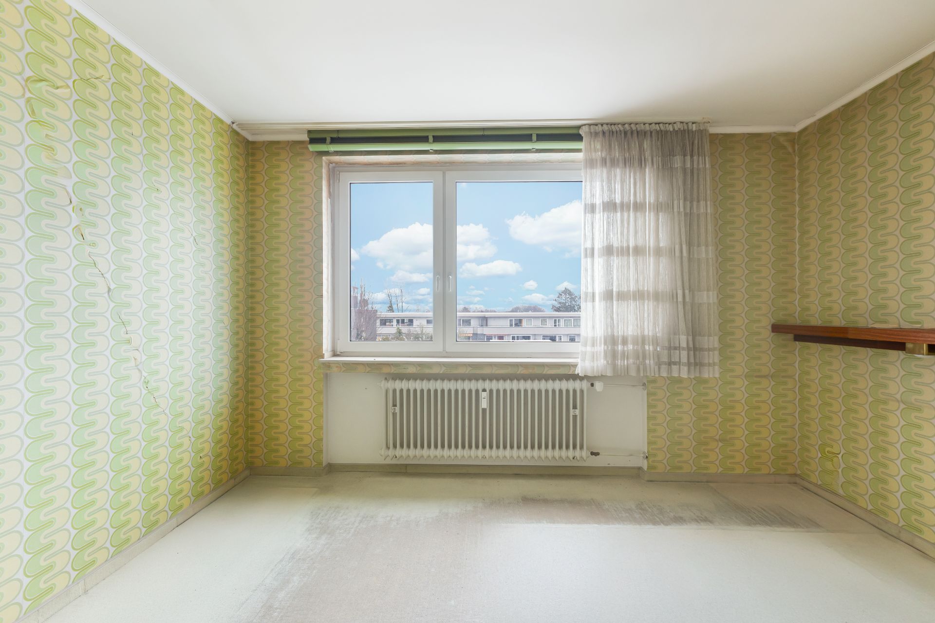Leerer Raum mit gemusterter Tapete, großem Fenster und Heizkörper darunter.