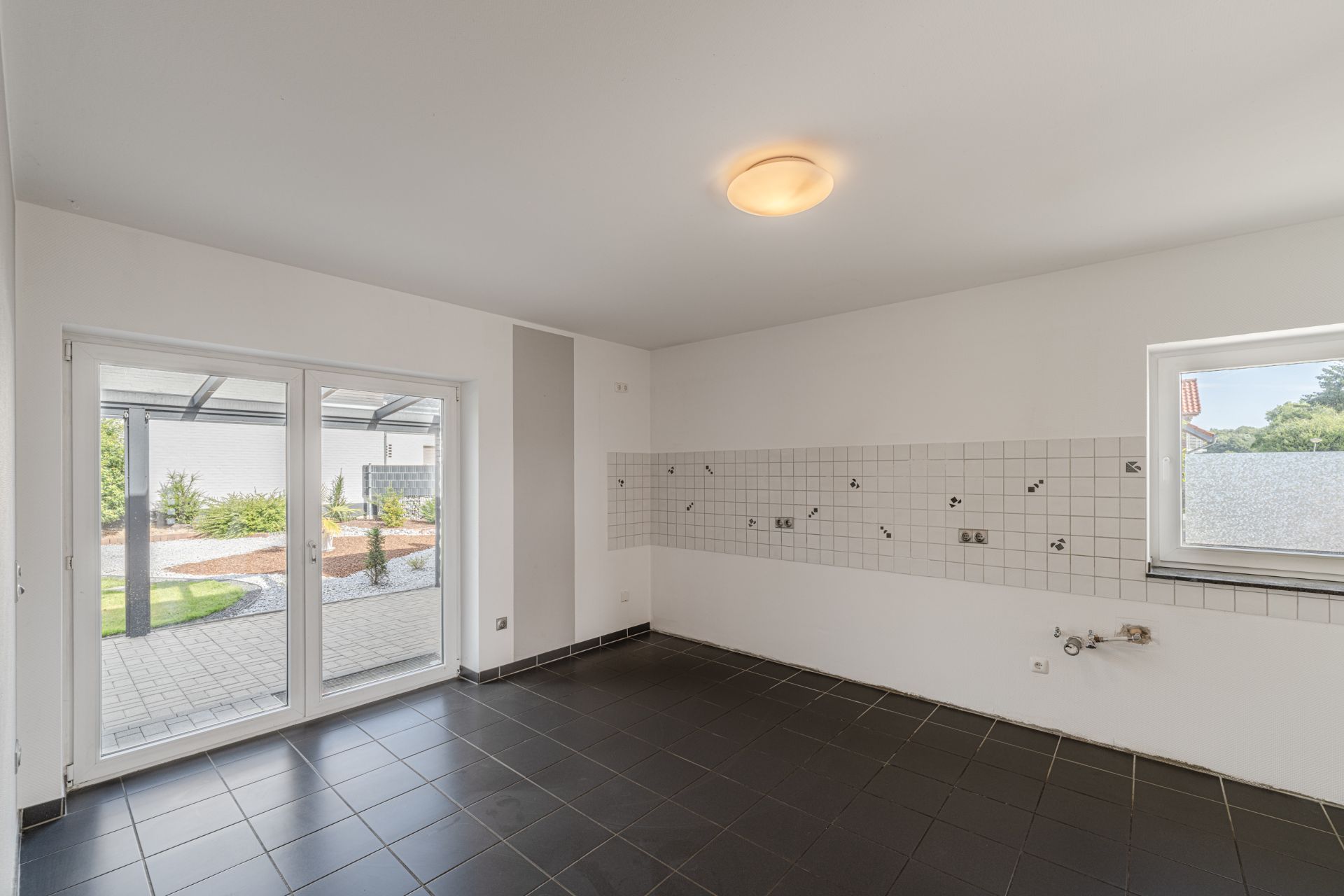 Leerer Raum mit großen Fenstern, Fliesenboden und gefliester Küchenwand.