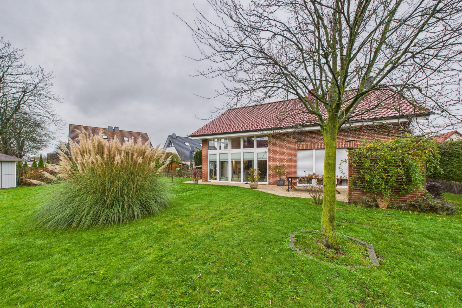 Backsteinwohnhaus mit rotem Dach, großer Garten, Baum und Ziergras im Vordergrund.