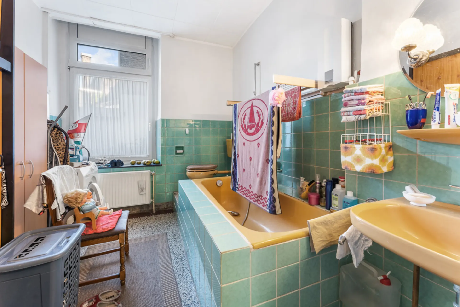 Badezimmer mit gelber Badewanne, Waschbecken, grünlichen Fliesen und verschiedenen Alltagsgegenständen.