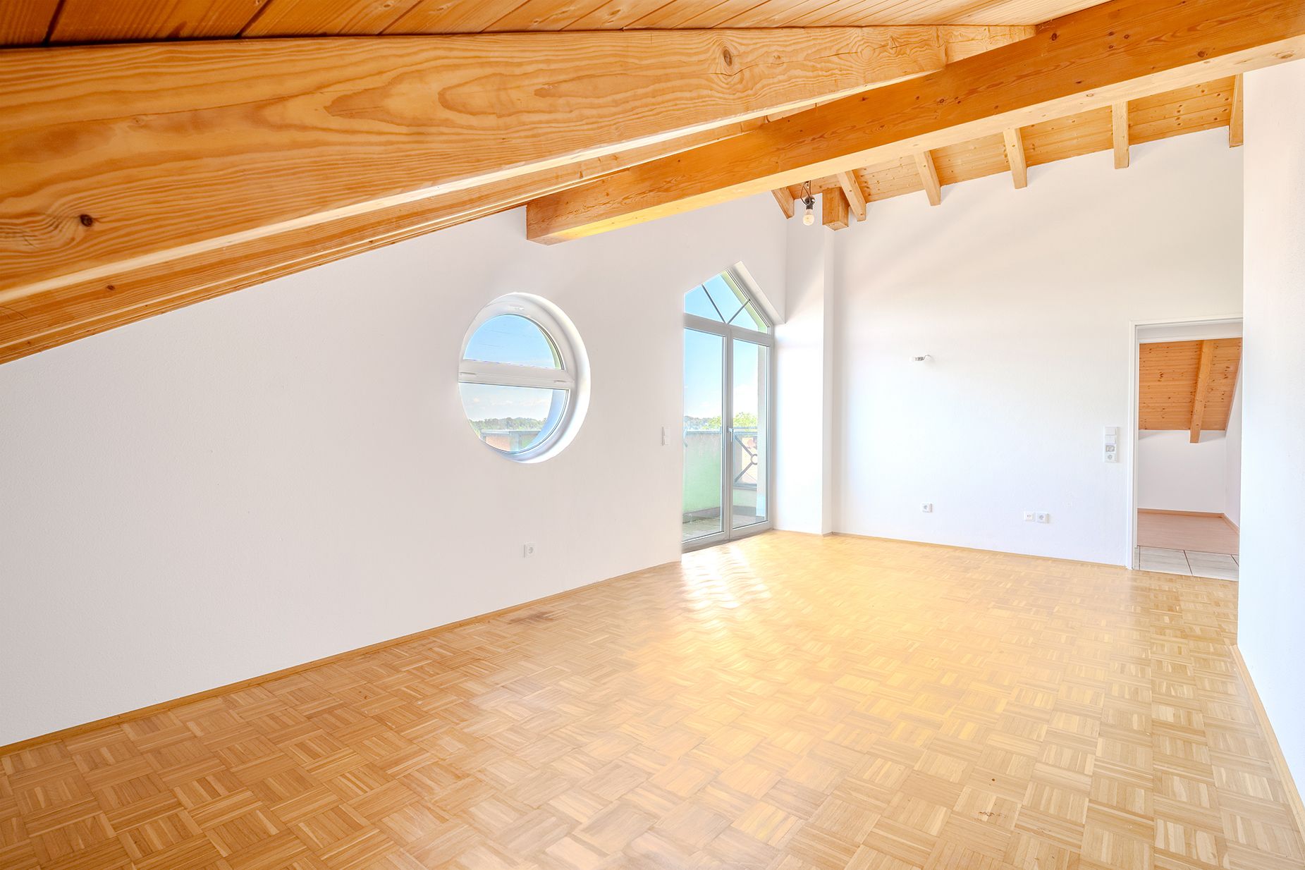 Helles Zimmer mit Holzdecke, Parkettboden, rundem Fenster und Zugang zu einem Balkon.