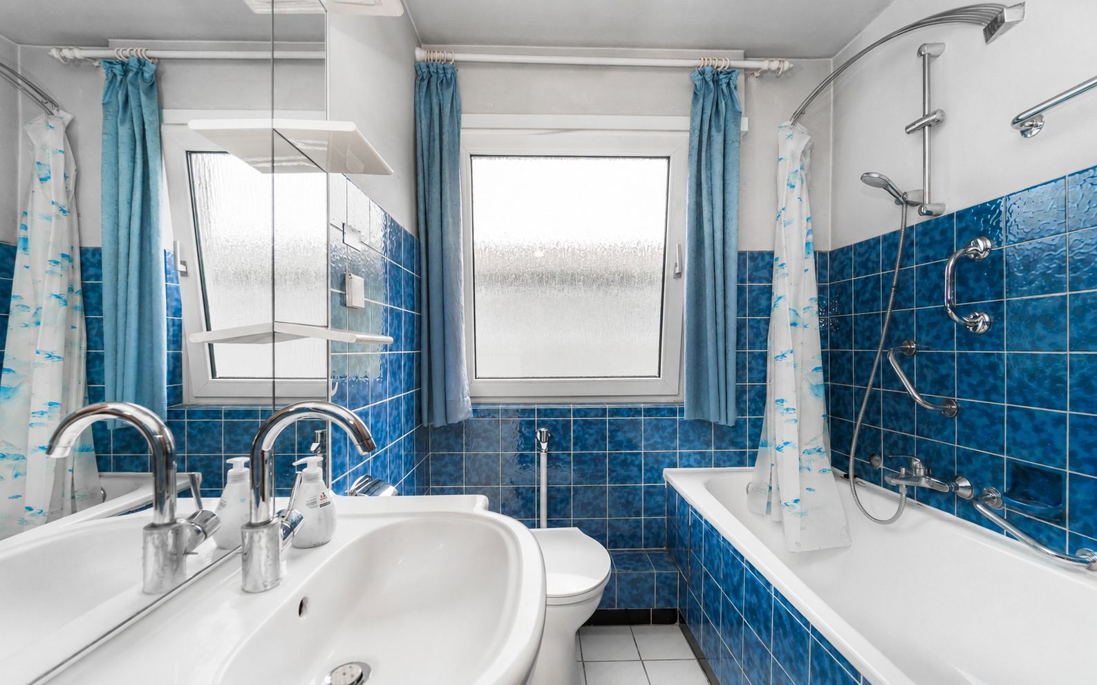 Badezimmer mit blauen Fliesen, Waschbecken, Spiegel, Toilette und Badewanne mit Duschvorhang.