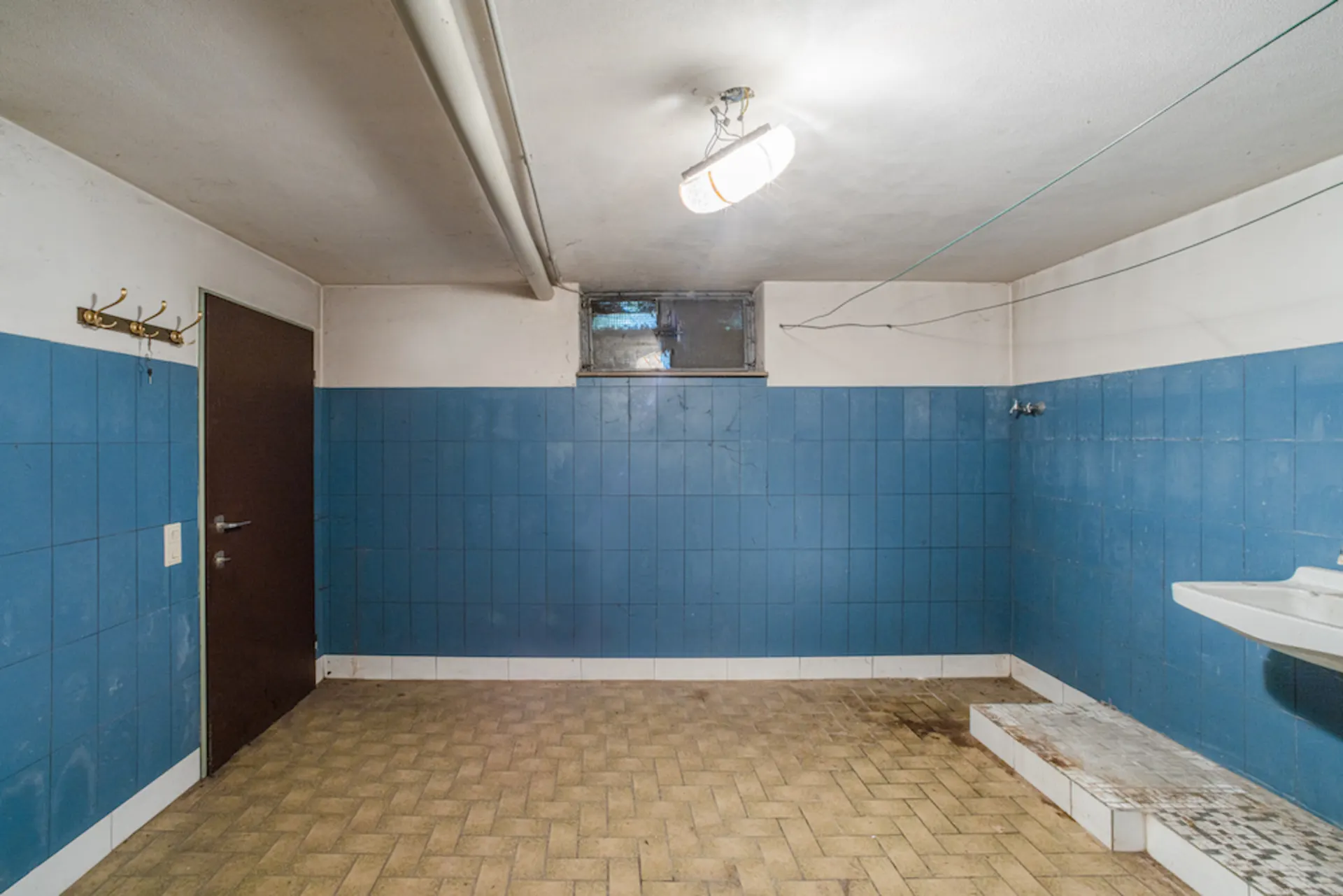 Kellerraum mit blauen Fliesen, brauner Tür, Waschbecken und Haken an der Wand.