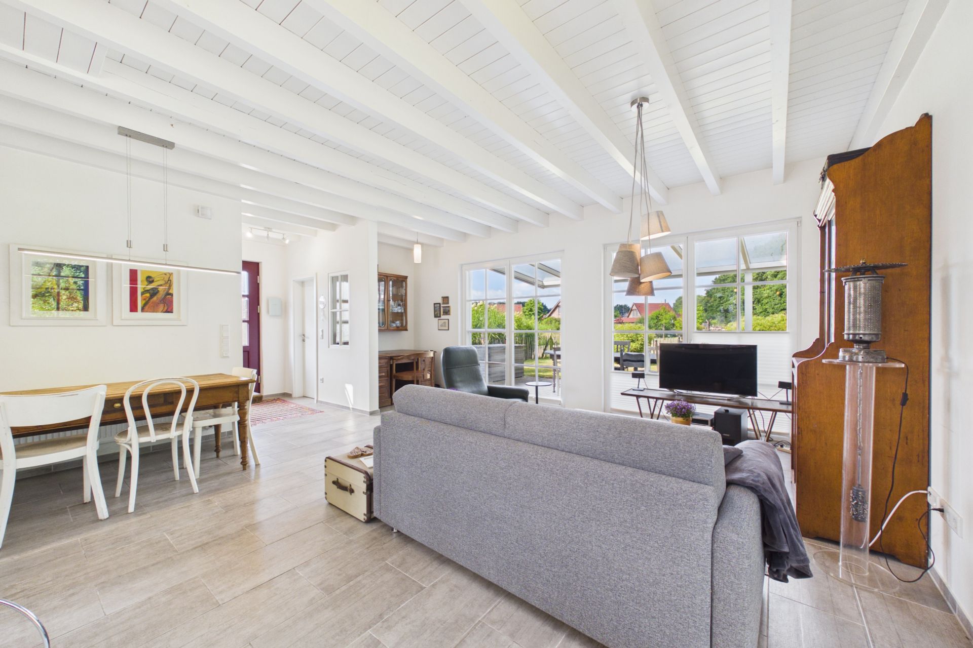 Helles Wohnzimmer mit grauem Sofa, Esstisch, Fernseher und großen Fenstern mit Blick ins Grüne.
