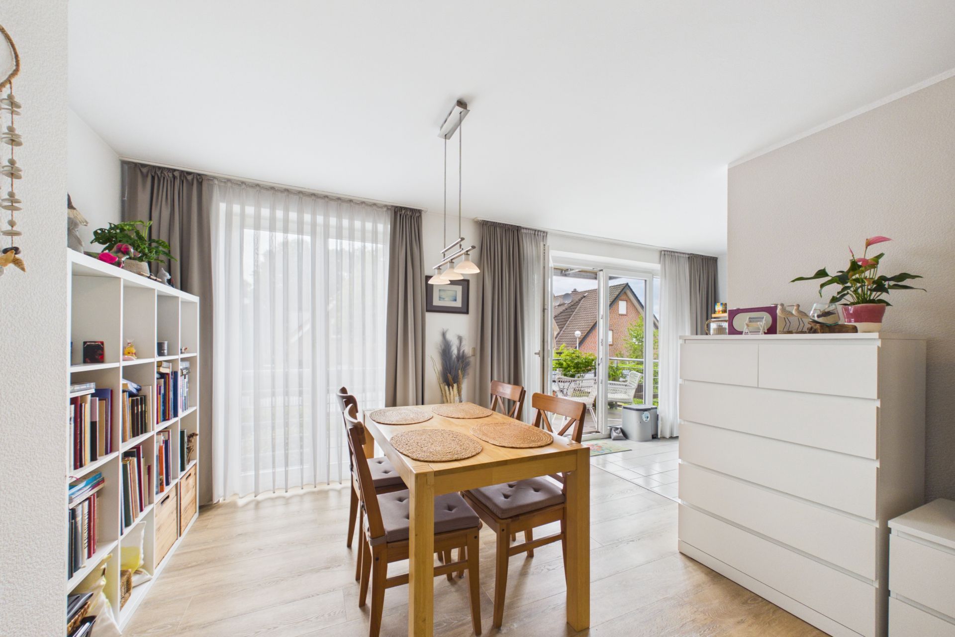 Essbereich mit Holztisch, vier Stühlen, Regal, Kommode und großen Fenstern mit Vorhängen.