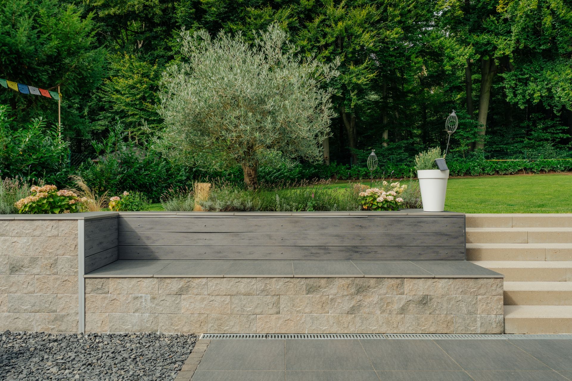 Garten mit steinerner Sitzbank, Stufen, Pflanzen und Bäumen im Hintergrund.