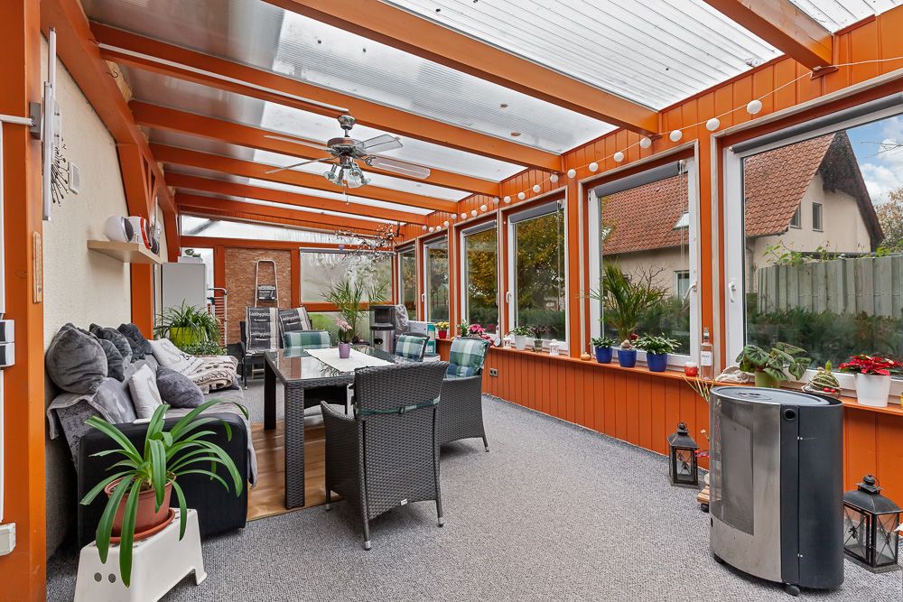 Heller Wintergarten mit Sofas, Esstisch, vielen Pflanzen und großen Fenstern.
