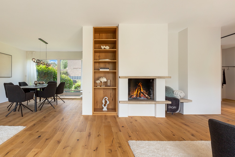 Modernes Wohnzimmer mit Kamin, Holzregal und Essbereich mit schwarzen Stühlen auf Holzboden.